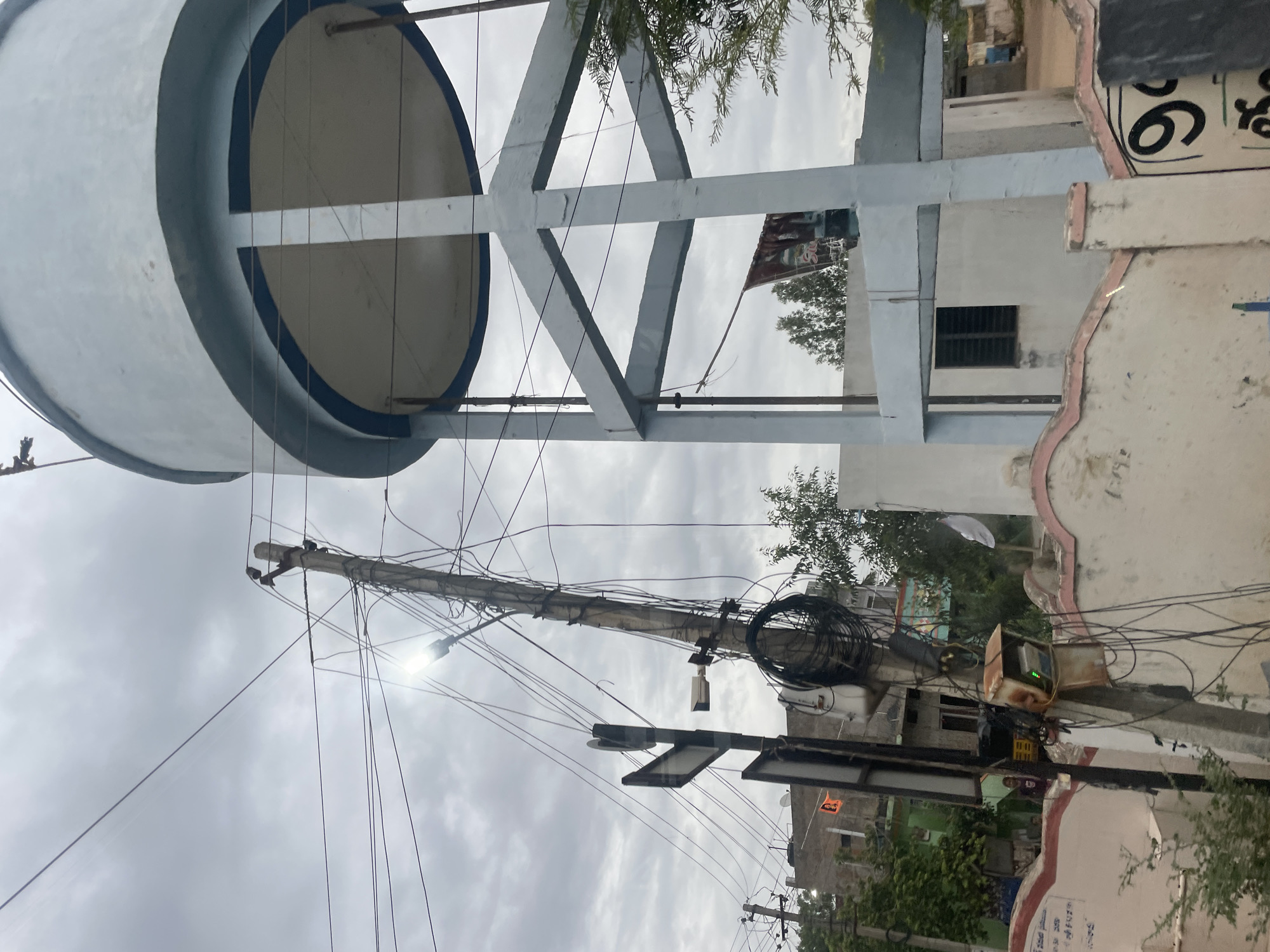 The image depicts a cloudy day in Bethamcherla, India, focusing on a water tank and a utility pole. The water tank, a large, light blue cylindrical structure, is supported by a grey, open-frame concrete base. The tank has a prominent dark blue band around its top and bottom edges. Below the tank, a white building with a window is visible.
To the left of the water tank is a concrete utility pole leaning precariously, heavily laden with a dense network of black electrical wires and cables. Attached to the pole are several items: a street light emitting a bright light, a security camera, and a signpost with two triangular signs, one of which appears to be a directional sign. Near the base of the pole, there is a rusty metal box with a green light, possibly an electrical meter or control unit, and coiled cables.
In the background, a densely packed residential area with multi-story buildings is visible. Some buildings have balconies and tiled roofs. A few trees and foliage are interspersed among the buildings. One building in the background has a flag with an orange symbol. A small, partially visible auto-rickshaw can be seen between two buildings.
In the foreground, a decorative white wall with pink trim lines the bottom of the image. On the wall, there are some inscriptions and large black numerals, including "9". A few green plants are growing in front of the wall. The overall impression is of a densely populated urban or semi-urban environment with visible infrastructure and signs of daily life.