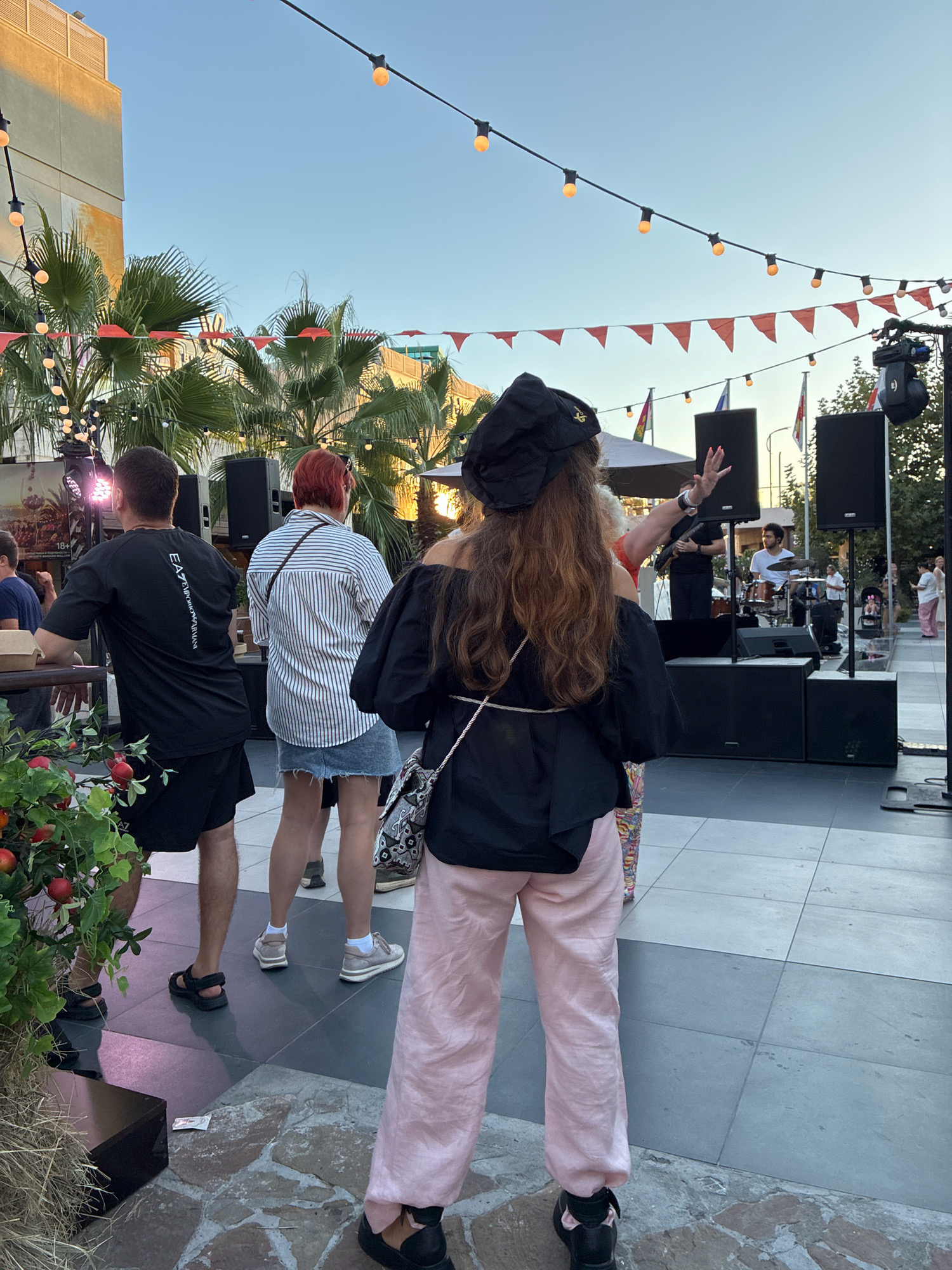 A group of people are gathered outdoors in what appears to be a plaza or pedestrian area, possibly in Sochi, Russia, during daylight hours. A band is performing on a raised stage in the background, with musical instruments and speakers visible. String lights and orange triangular flags are strung across the sky, suggesting a festive atmosphere or event. Several palm trees are also present, adding to a potentially warm-weather setting. People in the foreground are facing the stage, with one woman in a black hat and pink pants appearing to gesture towards the performance. Another person in a black t-shirt and shorts is standing to the left, and a woman with red hair in a striped shirt and denim skirt is to the right. The ground is paved with a mix of square tiles and irregular stones.
