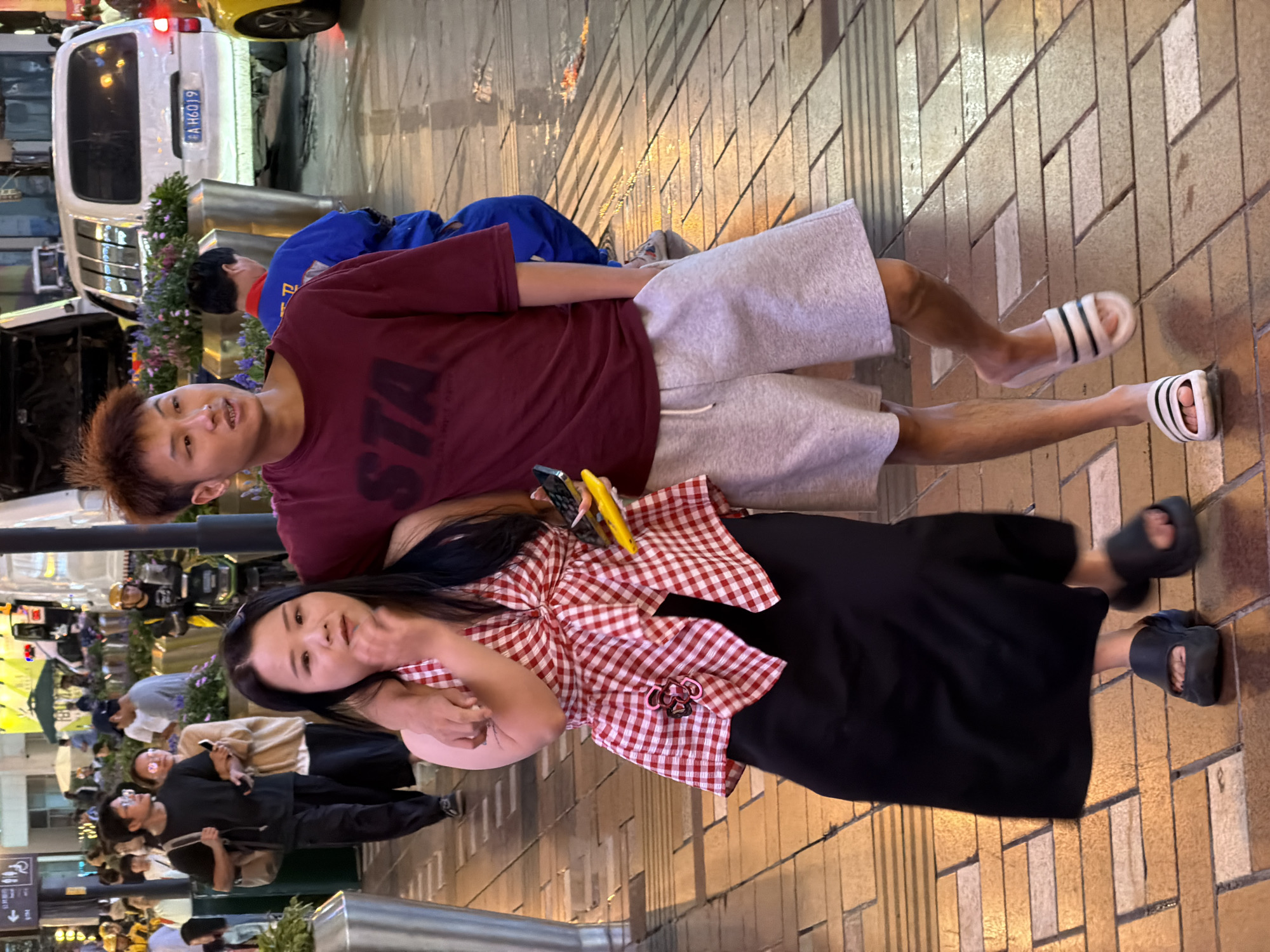 A couple walks down a tiled pedestrian street in Yuzhong District, China. The man in front is wearing a maroon t-shirt and light gray shorts, while the woman beside him has on a red and white checkered halter top and a long black skirt. The man has his arm around the woman's shoulders. In the background, a white van is parked and other pedestrians are visible. The street is wet, reflecting the lights from nearby buildings.