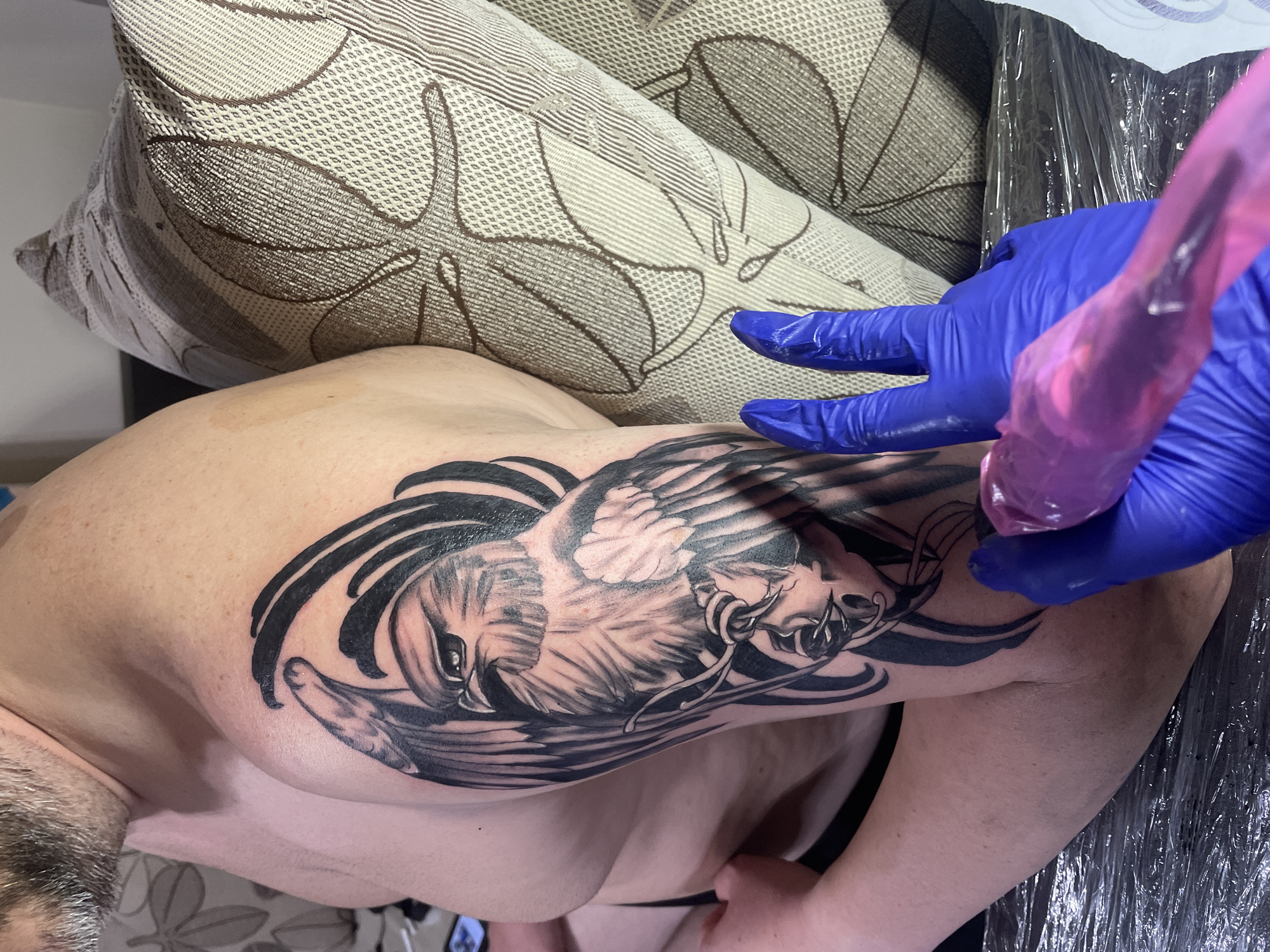 A person is receiving a tattoo of an eagle on their upper arm. The tattoo artist is wearing blue gloves and using a tattoo machine with a transparent pink grip. The person is lying on a couch with a decorative pillow nearby. The scene is likely taking place indoors, possibly at a tattoo parlor in Smolyan, Bulgaria.
