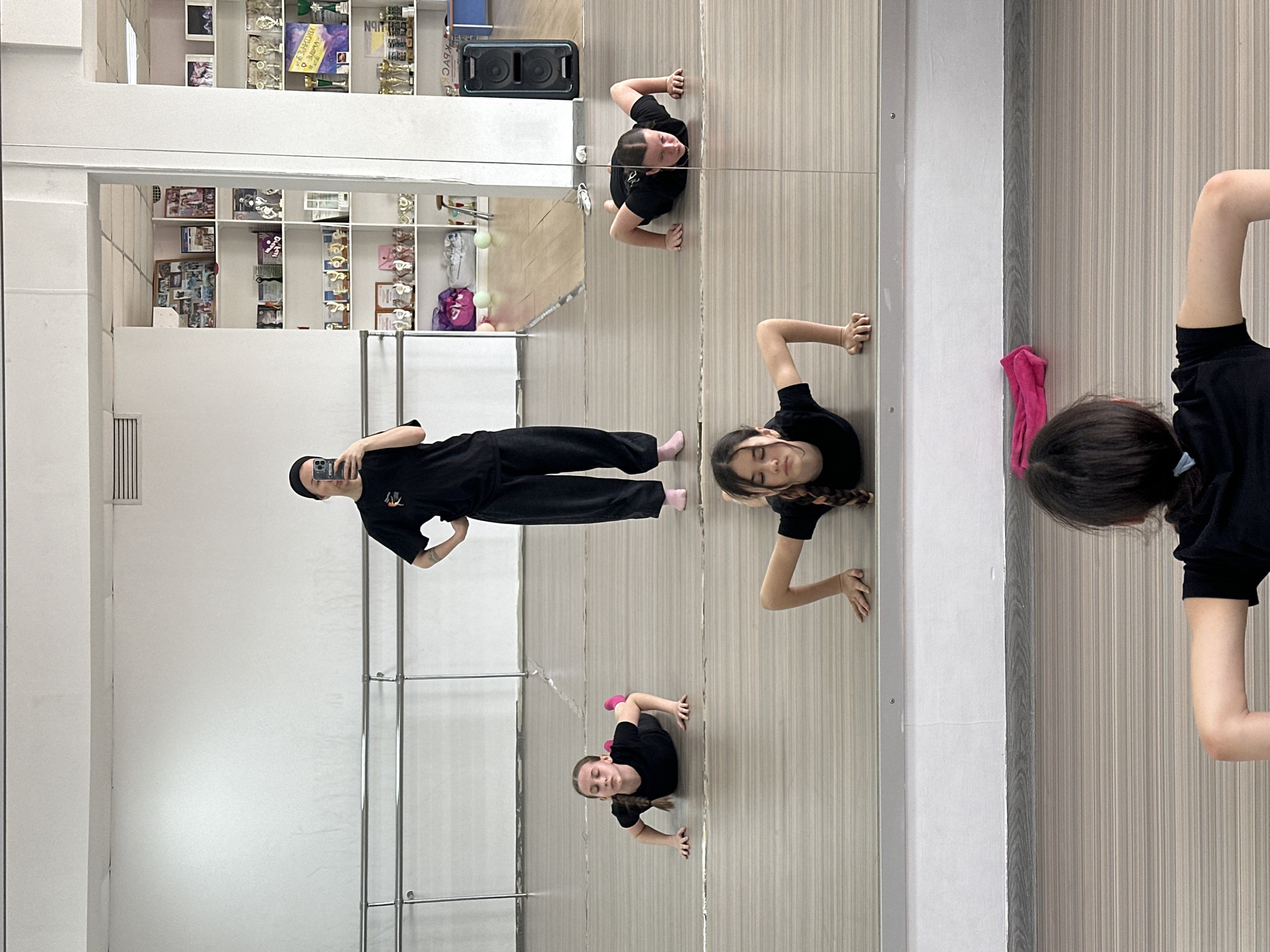 The image displays a mirror selfie taken in what appears to be a dance studio or gym in Votkinsk, Russia, rotated 90 degrees clockwise. This rotation presents the mirror's reflection as the primary vertical plane, with the actual floor and left wall appearing less prominently.
Reflected in the large mirror, a person, presumably the photographer, stands on the left, dressed in black pants, a black t-shirt, and a dark beanie, holding a smartphone to capture the image. Two young girls, also in black t-shirts and leggings, are positioned on what appears to be the "floor" within the reflection. One girl is in a plank or push-up stance, looking upwards, distinguished by pink socks. The second girl is closer, in a similar pose, with her face turned slightly away from the camera. A third girl's head and upper body are partially visible on the far right, also in a comparable pose, wearing a black t-shirt.
The background, seen in the mirror behind the photographer's reflection, consists of white shelves filled with numerous trophies, awards, and what seem to be dance shoes. A black speaker rests on a shelf. Visible text includes "DANCE" on a purple poster and "VOTKINS" on an award or sign, aligning with the provided location context. A pink towel is draped over a vertical divider or bar on the right. The studio's actual floor, visible on the left, features a light wood plank pattern, and chrome support bars are also reflected. The indoor lighting is bright and uniform, highlighting the subjects' focused expressions.