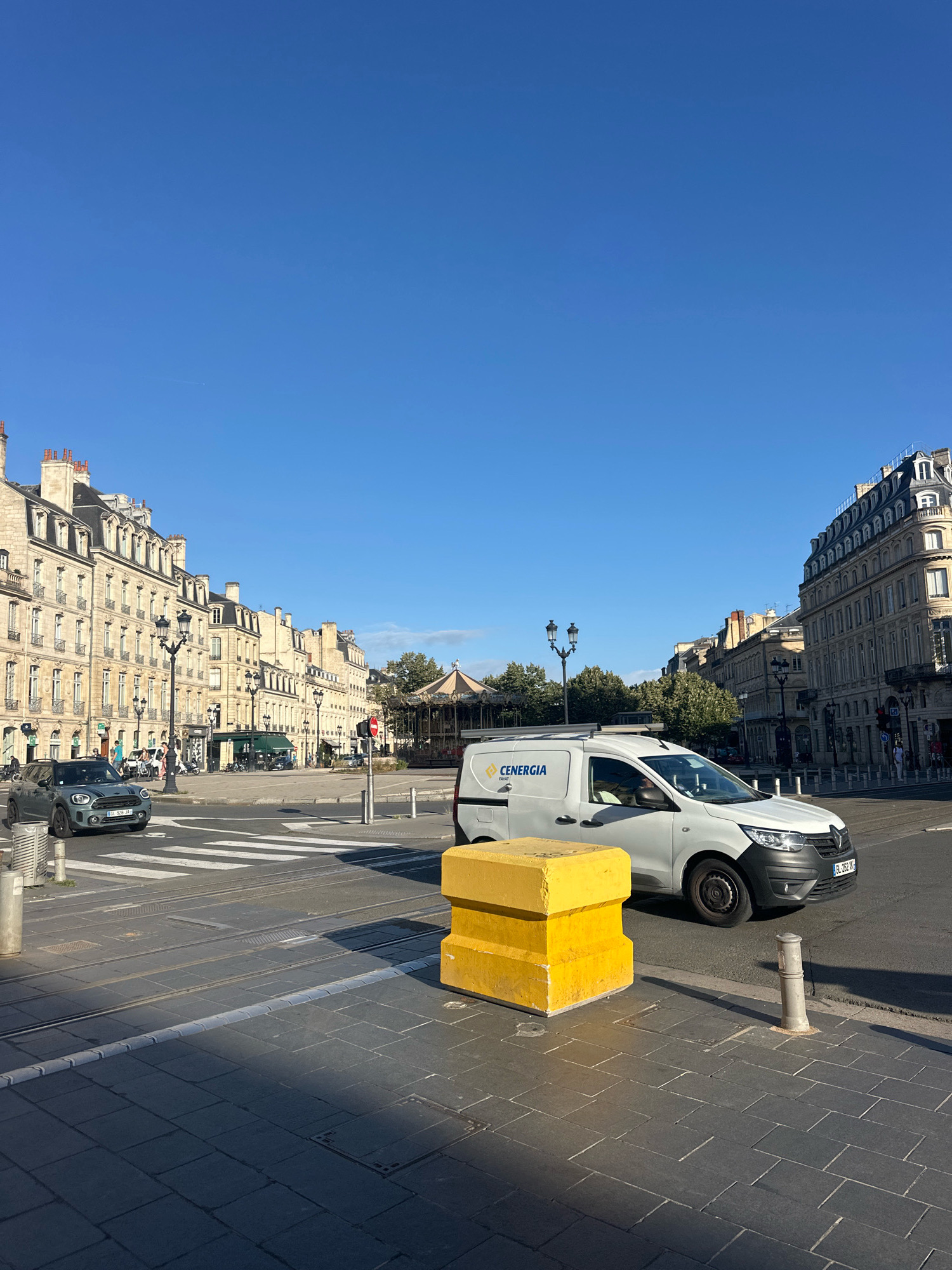 This is a street-level, daytime, medium shot of a wide intersection in Bordeaux, France. The sky is clear and blue. On the left, a row of ornate, multi-story buildings with Mansard roofs lines the street. A green Mini Cooper is stopped at a crosswalk. In the center of the intersection, there is a carousel. On the right, more ornate buildings with Mansard roofs are visible. A white van with the logo "CENERIA" is driving through the intersection. In the foreground, a yellow concrete barrier is placed on the sidewalk, and lampposts and bollards are visible. Tram tracks are present on the road.