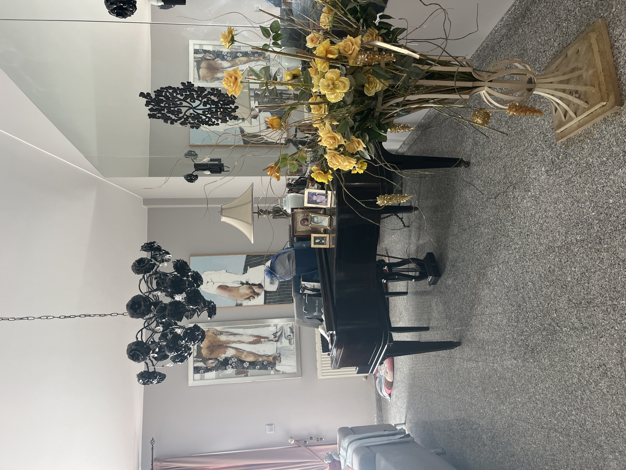 The image shows an interior scene, likely a living room or music room, in Parekklisia Community, Cyprus. Dominating the foreground and center is a dark, polished grand piano. To the right of the piano, a tall, ornate vase holds a large arrangement of yellow roses and decorative branches, some adorned with gold accents. A lamp with a cream-colored shade sits on the piano's surface, next to a collection of framed photographs.
In the background, two large, framed paintings hang on the wall. The painting on the left depicts the lower body and legs of a person wearing patterned socks and shoes, against a black and white floral background. The painting on the right features a nude torso and foot. Above the piano, a dramatic black chandelier with rose-shaped accents hangs from the ceiling.
A large mirror panel reflects the room, showing additional decorative elements like wall sconces and a filigree wall art piece. To the left of the piano, a portion of a grey sofa is visible, draped with a light blue throw blanket. A pink and striped floor cushion lies on the speckled grey carpet. The walls are painted a neutral grey, and pink curtains hang from a rod on the left side of the frame. The environment appears to be a well-decorated indoor space.