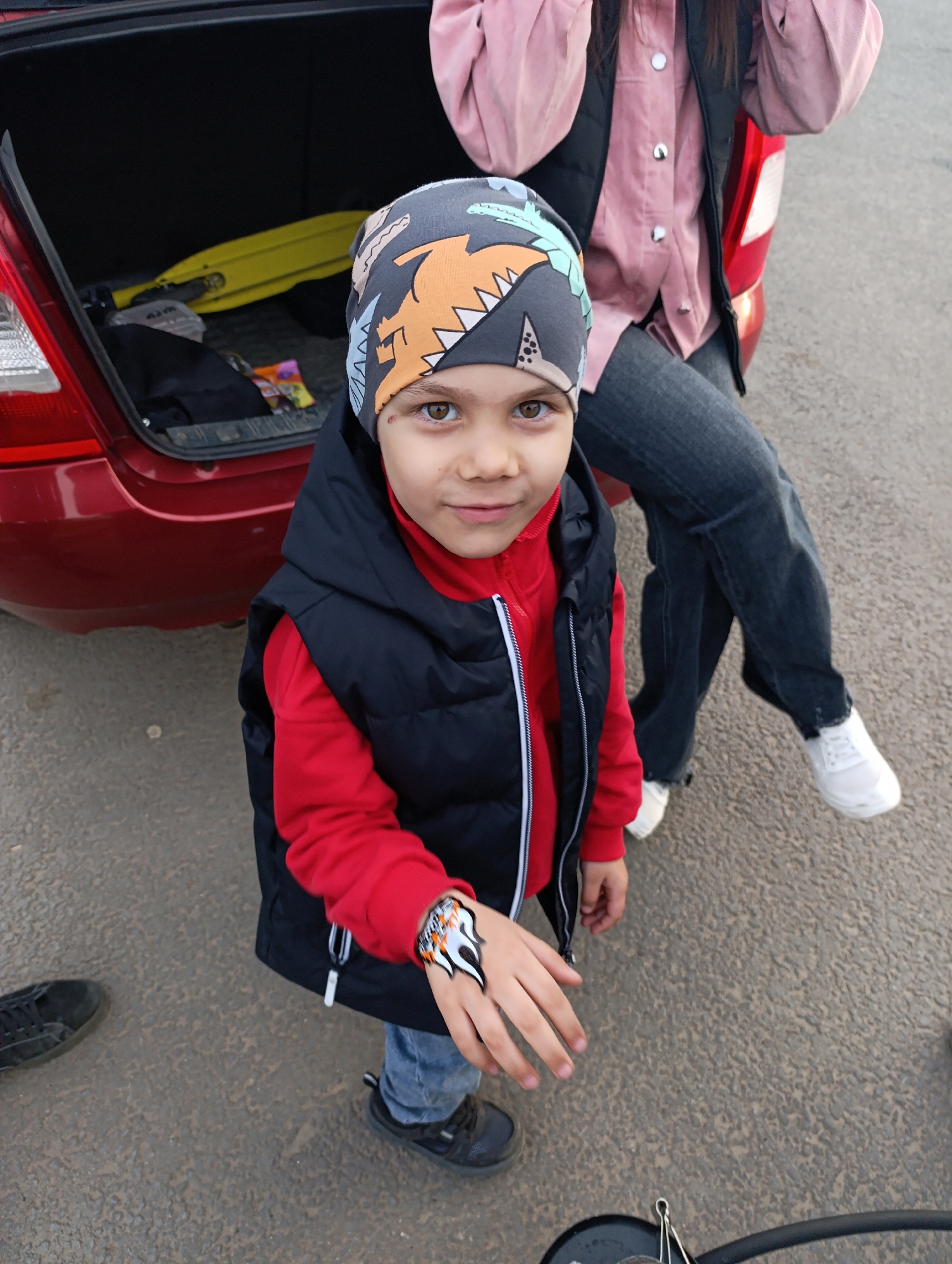 A young child is the focal point of this eye-level full shot, standing outdoors in front of the open trunk of a red car. The child is wearing a gray beanie with dinosaur designs, a red hooded sweatshirt, a black vest, blue jeans, and black athletic shoes. A temporary tattoo adorns the child's outstretched right hand.
Behind the child, partially visible, is a person with their legs dangling off the trunk of the red car. The person is wearing a pink button-down shirt, a black vest, blue jeans, and white sneakers. The trunk of the red car is open, revealing a yellow scooter and other unidentified items inside.
The scene is set on an asphalt surface, likely a parking lot or roadside. A black shoe and a black object, possibly a car part or tool, are visible on the ground near the bottom of the frame. The specific location in the city of Unknown, Unknown, is not discernable from the visual information provided.