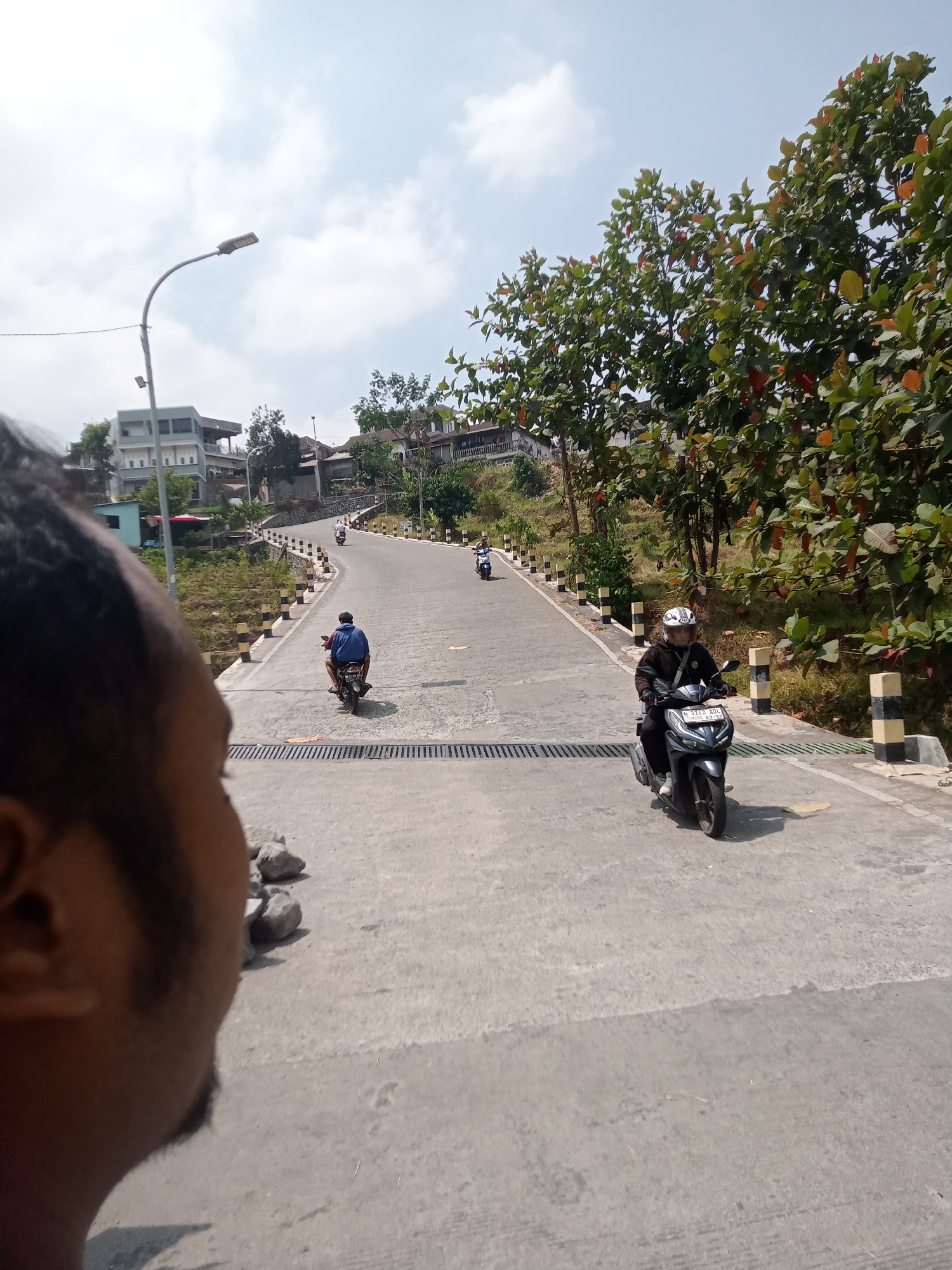 The image captures a view of a sloping road in Batu, Indonesia, with several motorbikes navigating its incline. In the foreground, the profile of a person's head and shoulder is visible on the left, obscuring a portion of the scene. The road itself is paved and marked with yellow and black striped bollards along the sides. Trees with green and reddish-brown leaves line the upper portion of the road, casting shadows on the pavement. Buildings, likely residences or small businesses, are situated on the hillside to the left and in the background. A street lamp is also visible on the left side of the road. Four motorbikes are present in the frame: one in the foreground being ridden by a person wearing a white helmet and dark clothing, another with a rider in a blue shirt further up the road, and two more motorbikes positioned even higher up the slope, their riders and vehicles less distinct. The sky above is partly cloudy. The scene appears to be a typical day on a road within a residential or semi-rural area of Batu.