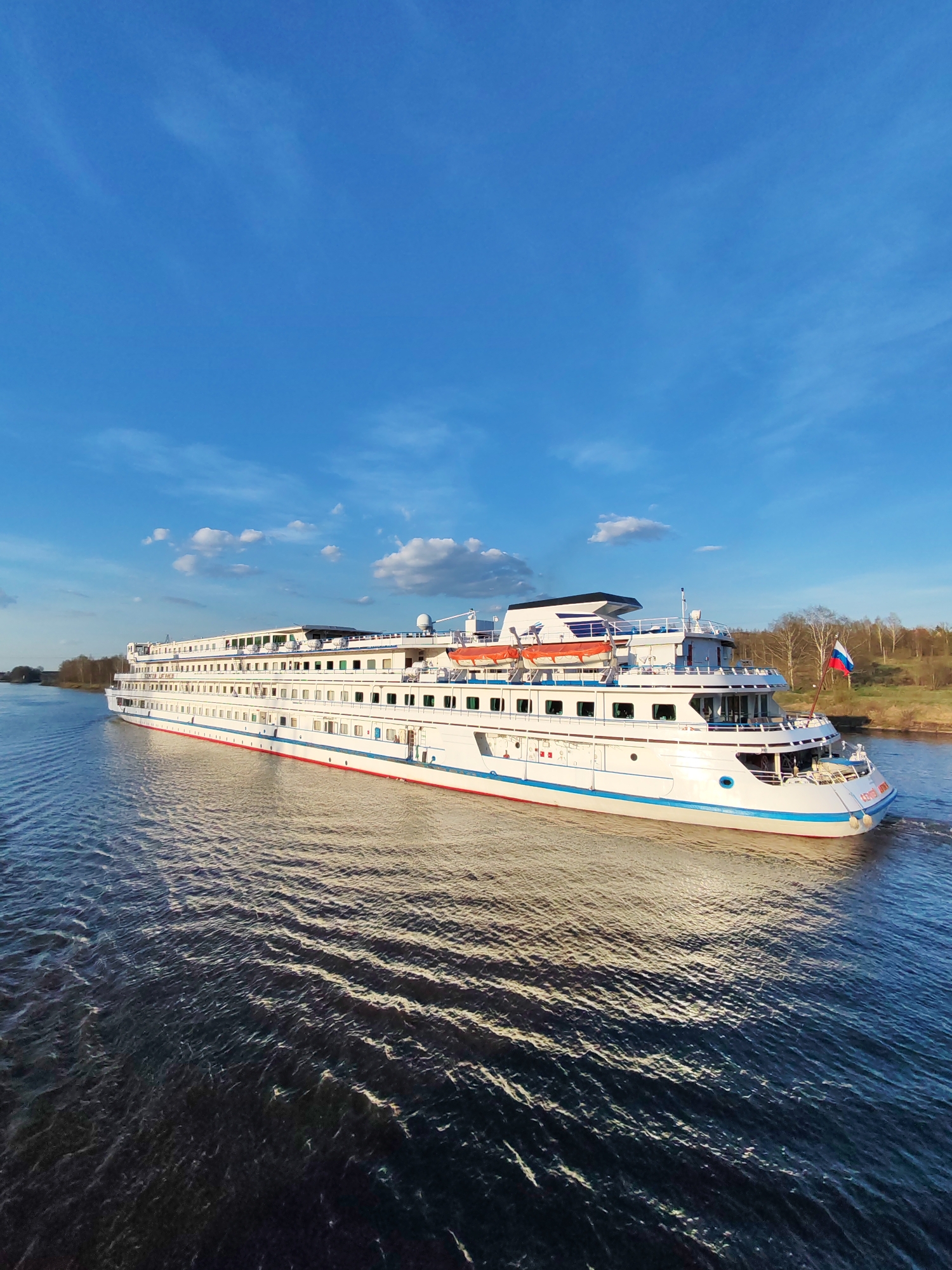The image shows a large white river cruise ship moving on a river near Базарово, Russia, under a clear blue sky with scattered clouds. The ship has multiple decks with rows of windows, and a Russian flag is visible at the stern. The water is dark blue and slightly rippled. Along the riverbank, there are trees and vegetation. The overall scene is bright and appears to be taken during daylight hours.