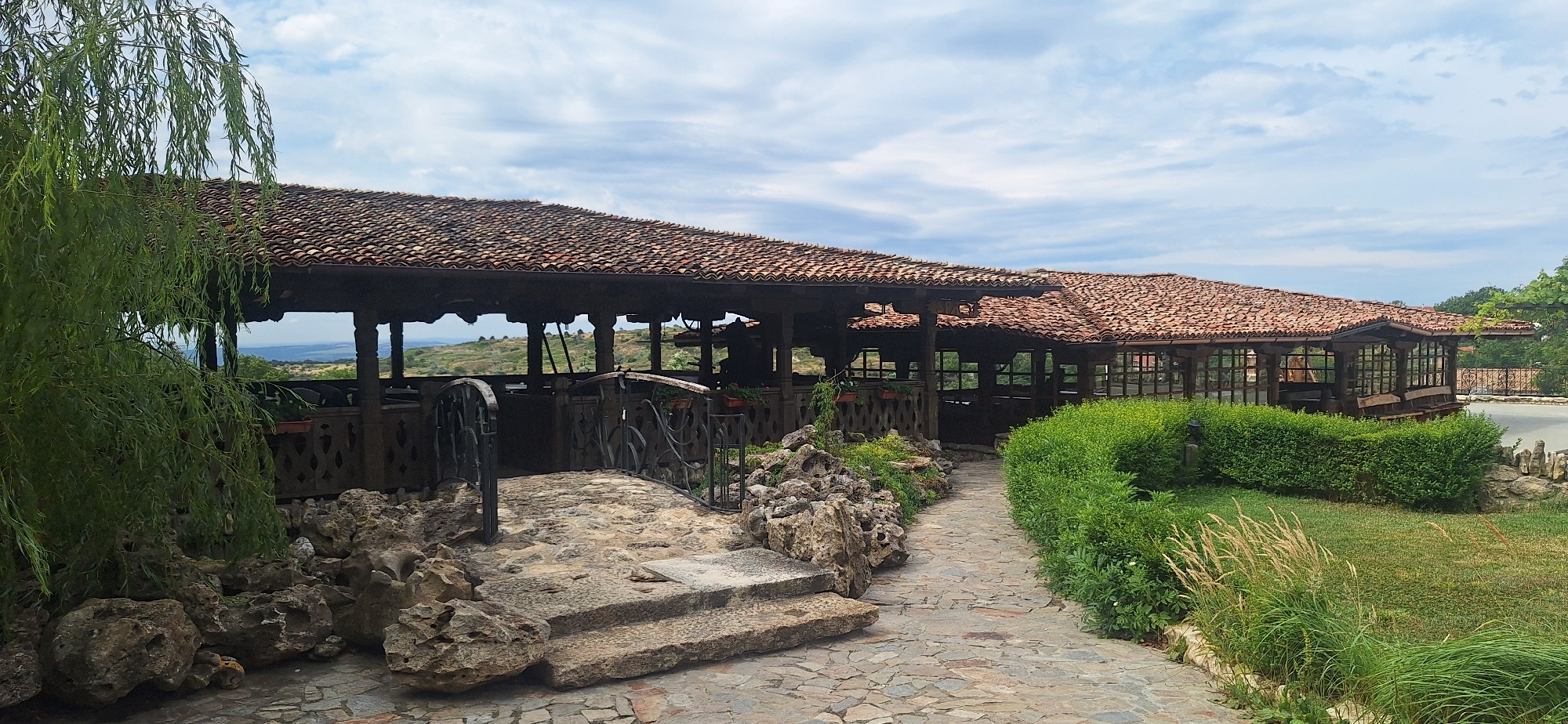 The image shows a restaurant with a traditional tiled roof, nestled in a lush green environment. The restaurant has an open-air seating area supported by wooden pillars, and a section enclosed with wooden frames and glass. A stone pathway leads to the restaurant, bordered by green bushes and a natural rock formation. The overall scene suggests a serene dining experience with a scenic view, likely in a rural setting within Kalimantsi, Bulgaria, given the context of the image. There are no people visible in the image. The sky is partly cloudy, indicating daytime.