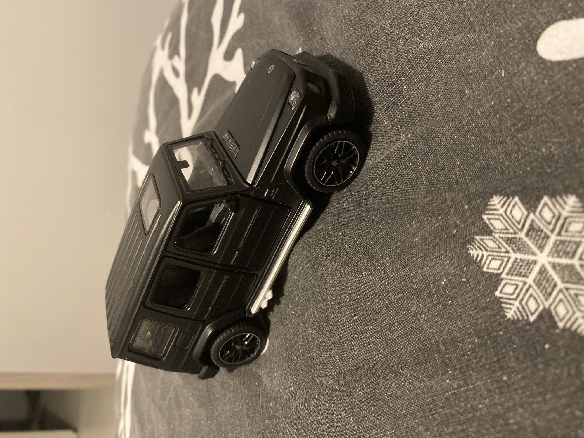 A black Mercedes-Benz G-Wagon toy car is positioned on a dark grey fabric surface, which has a pattern of white snowflakes and abstract shapes. The toy car is angled slightly, with its front right side closer to the viewer. It is a detailed replica, featuring visible headlights, grille, side mirrors, door handles, windows, and wheels. The car has a sunroof. The fabric surface appears to be a blanket or bedspread. The background is blurred and suggests an indoor setting with a light-colored wall and a doorway on the left. The lighting is artificial and casts soft shadows. There is no indication of the specific location within the city of Łódź, Poland.