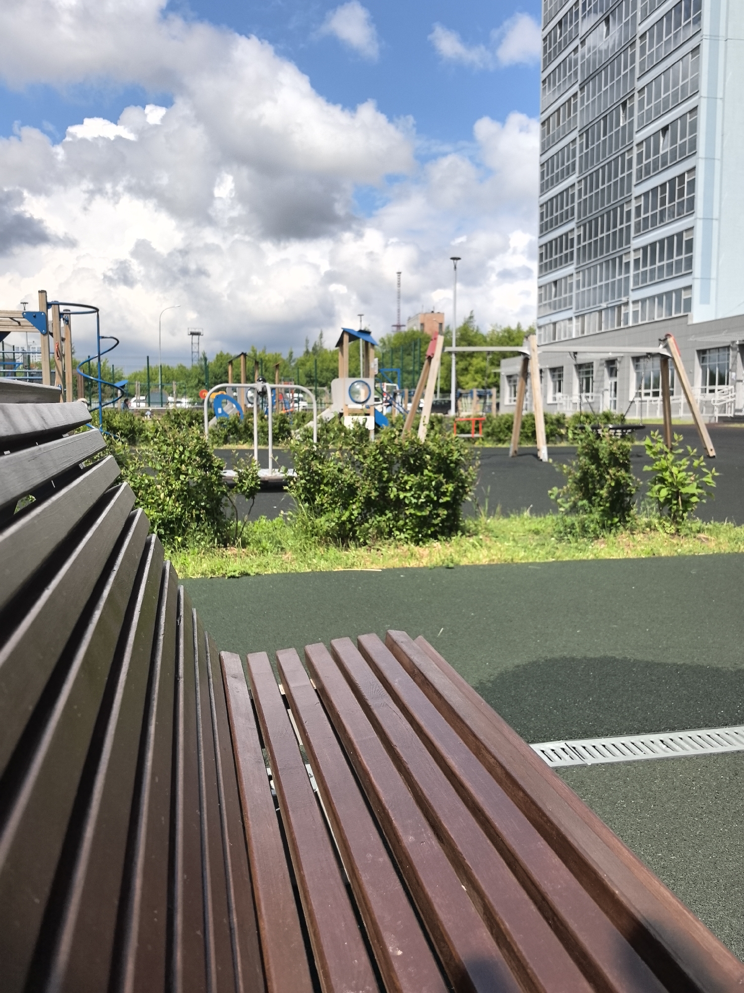 The image captures a modern urban playground scene on a bright, partially cloudy day in Perm, Russia. The perspective is from a low angle, placing a dark brown wooden park bench prominently in the foreground, extending from the bottom left to the lower right. The slats of the bench's backrest are sharply in focus on the left, while the seat recedes towards the right.
Beyond the bench, the midground reveals a well-maintained children's playground with a dark green, rubberized safety surface. Various colorful play structures are visible, including a blue and white climbing frame with a spiral element, a white and blue merry-go-round, and a wooden swing set with two swings. Small, lush green bushes are strategically planted around the play equipment, breaking up the open space.
In the background on the right, a tall, light-colored modern residential building with numerous windows dominates the skyline. Its design suggests contemporary architecture. Further in the background on the left, a line of green trees indicates a park or natural area bordering the playground, with a tall, slender communication tower visible in the distance above the trees. A smaller building with a reddish roof is partially seen behind the trees and playground structures.
The sky above is a vibrant blue with large, white cumulus clouds, suggesting fair weather. The bright light indicates it is daytime, likely mid-morning or afternoon. No people are present, lending a quiet and serene atmosphere to the scene. A metal drain grate is visible on the rubberized ground near the right side of the bench. There is no visible text in the image.