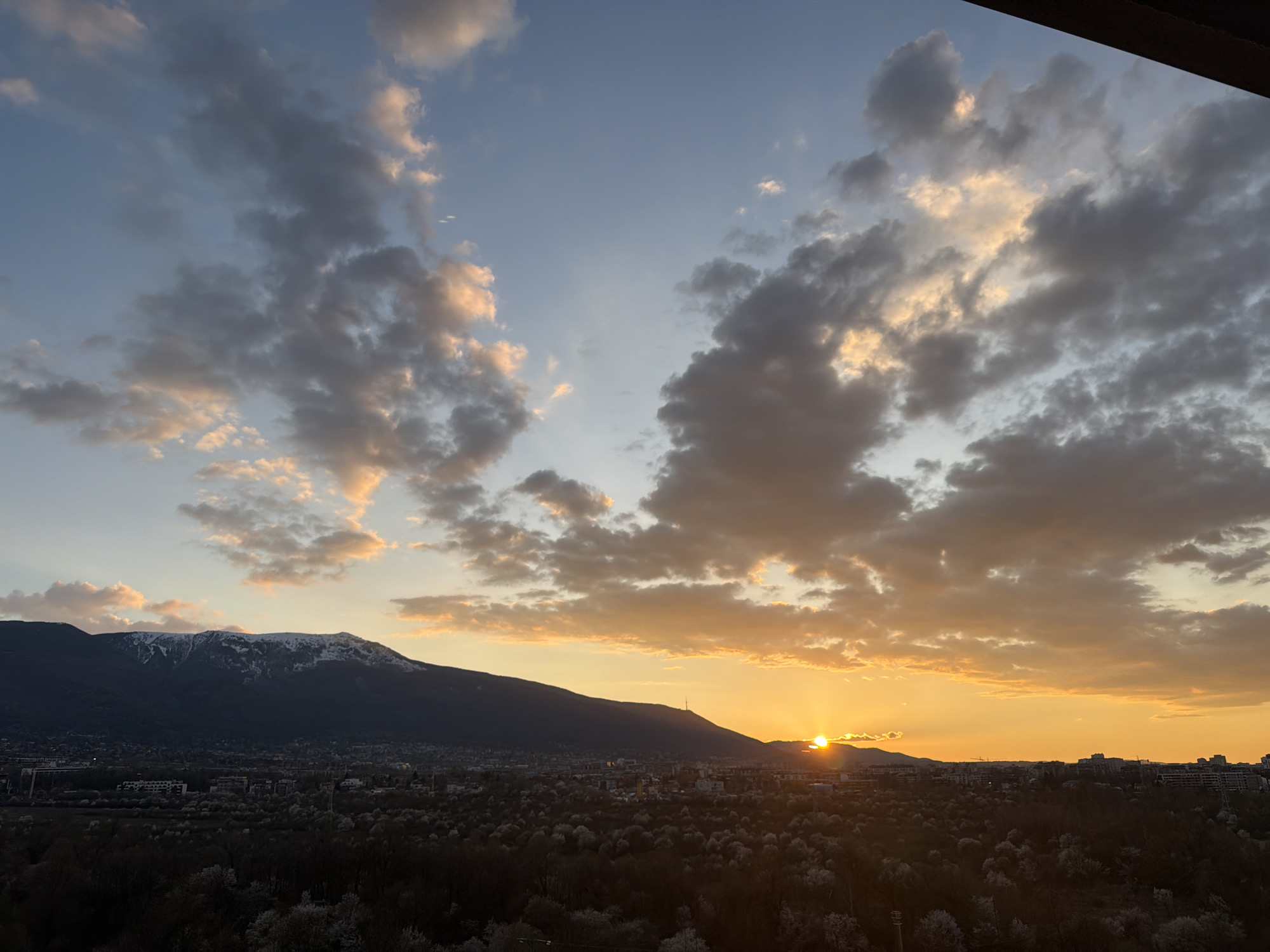 The image depicts a landscape at sunset, likely in Sofia, Bulgaria. A mountain range dominates the left side of the image, with some snow visible on the peaks. Below the mountains is a densely populated area with buildings and trees. The sun is setting behind another mountain range on the right, casting a warm orange glow across the sky and clouds. The sky is a mix of blue and orange, with scattered clouds. The overall scene suggests a tranquil and scenic view.