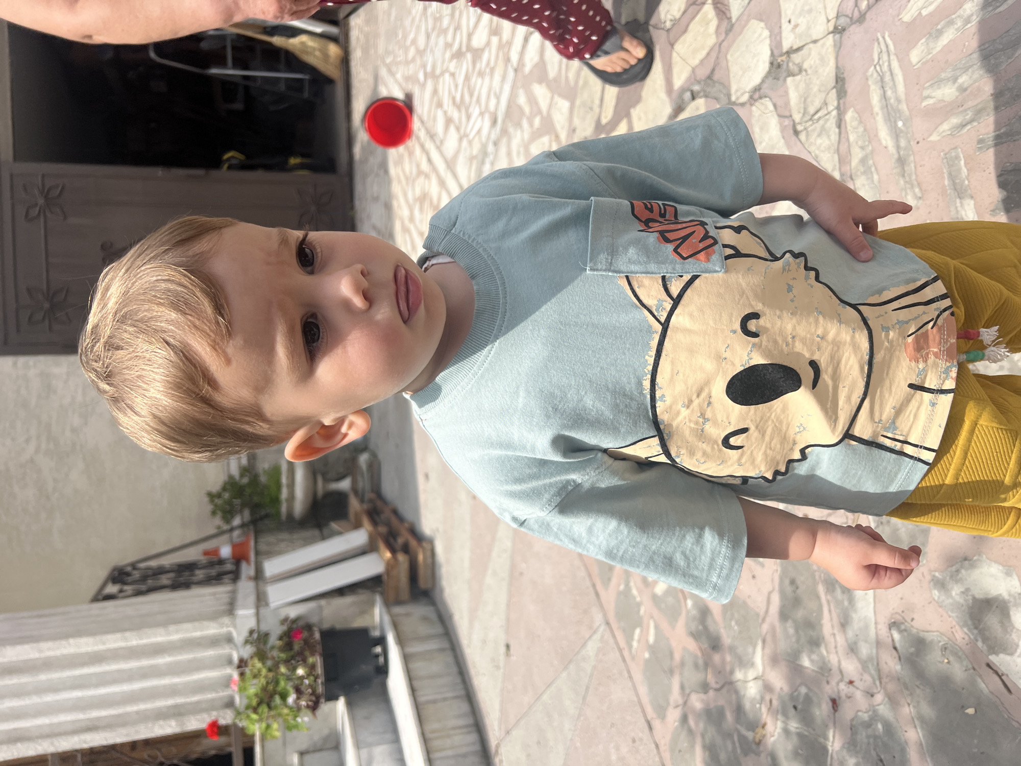 A young boy stands outside on a paved patio. He has light brown hair and is wearing a light blue t-shirt with a koala graphic and yellow pants. His mouth is slightly downturned, and his tongue is sticking out slightly. In the background, there is a large door, a red bucket, a broom, and potted plants. The lighting suggests it is daytime and sunny.