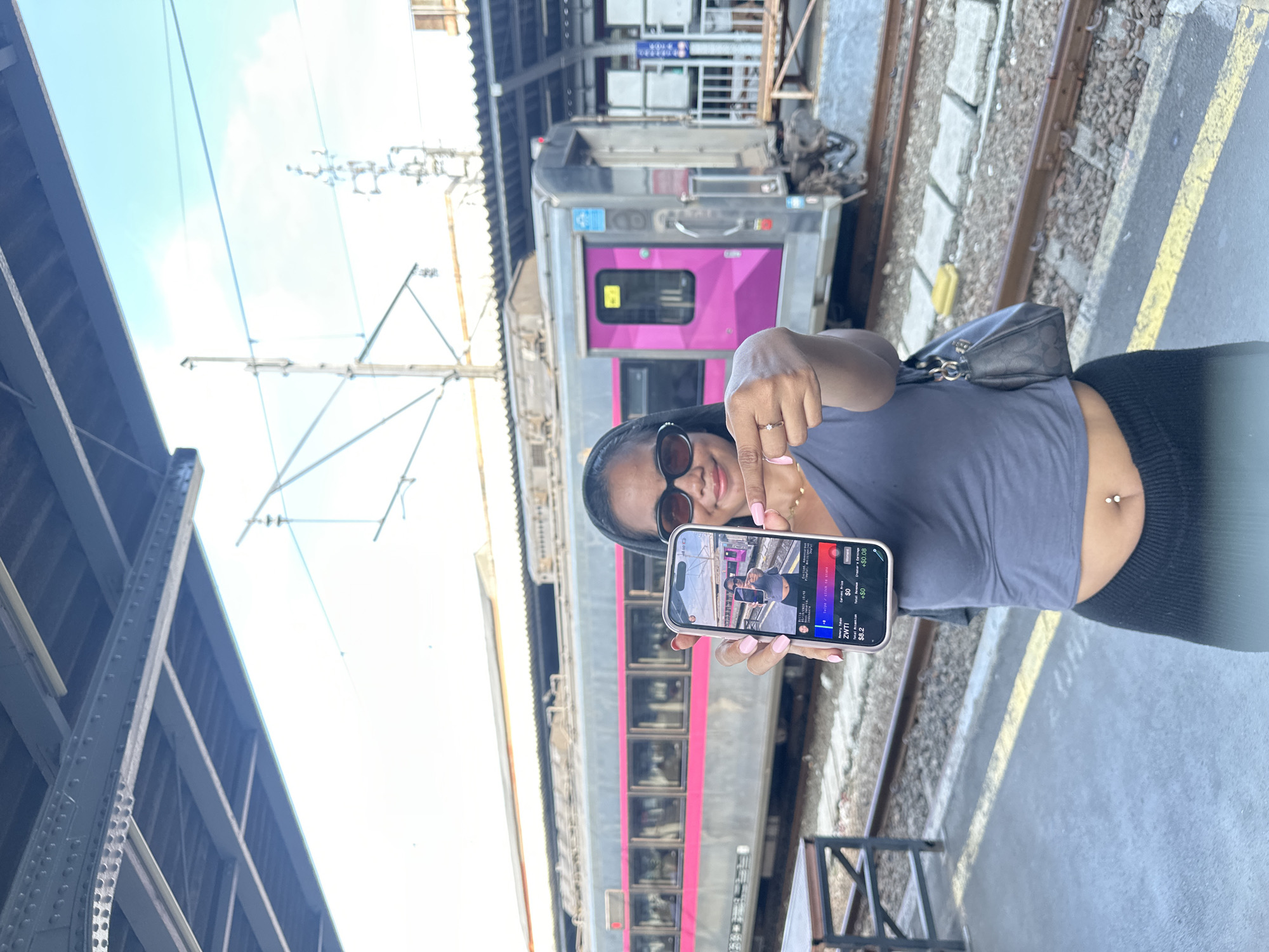 The image shows a person holding a smartphone, pointing at the screen with their index finger. The smartphone displays a photo of the same person holding a smartphone, creating a recursive effect. The person has dark hair and is wearing sunglasses, a gray top, and black bottoms. A silver train with pink accents is in the background. The setting appears to be a train station platform. The OCR content on the smartphone screen reads "so Swipe/ click to state ZWTI $8.2 +$0 +$0.08". There is no indication of the city of West Jakarta, Indonesia, present in the image.