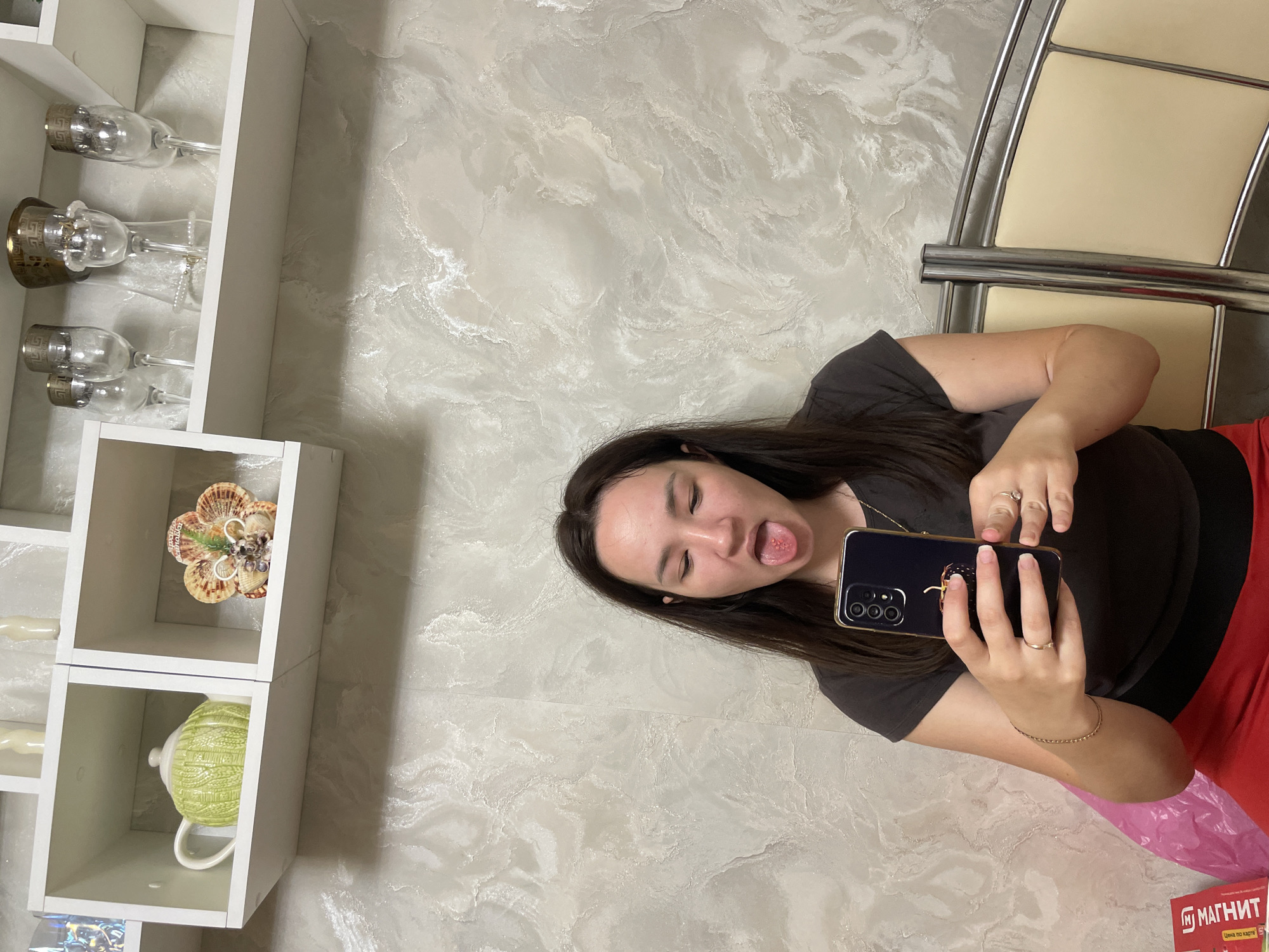 A young woman with dark hair is taking a selfie with her phone. She is sticking her tongue out, which has several small red sores on it. She is wearing a dark grey t-shirt and red pants. She is sitting on a bed with a light cream colored headboard. Behind her is a textured wall. On a shelf above her, there are several decorative items, including a green teapot and a shell ornament. The context of Laishevo, Russia is not inferable from the image.