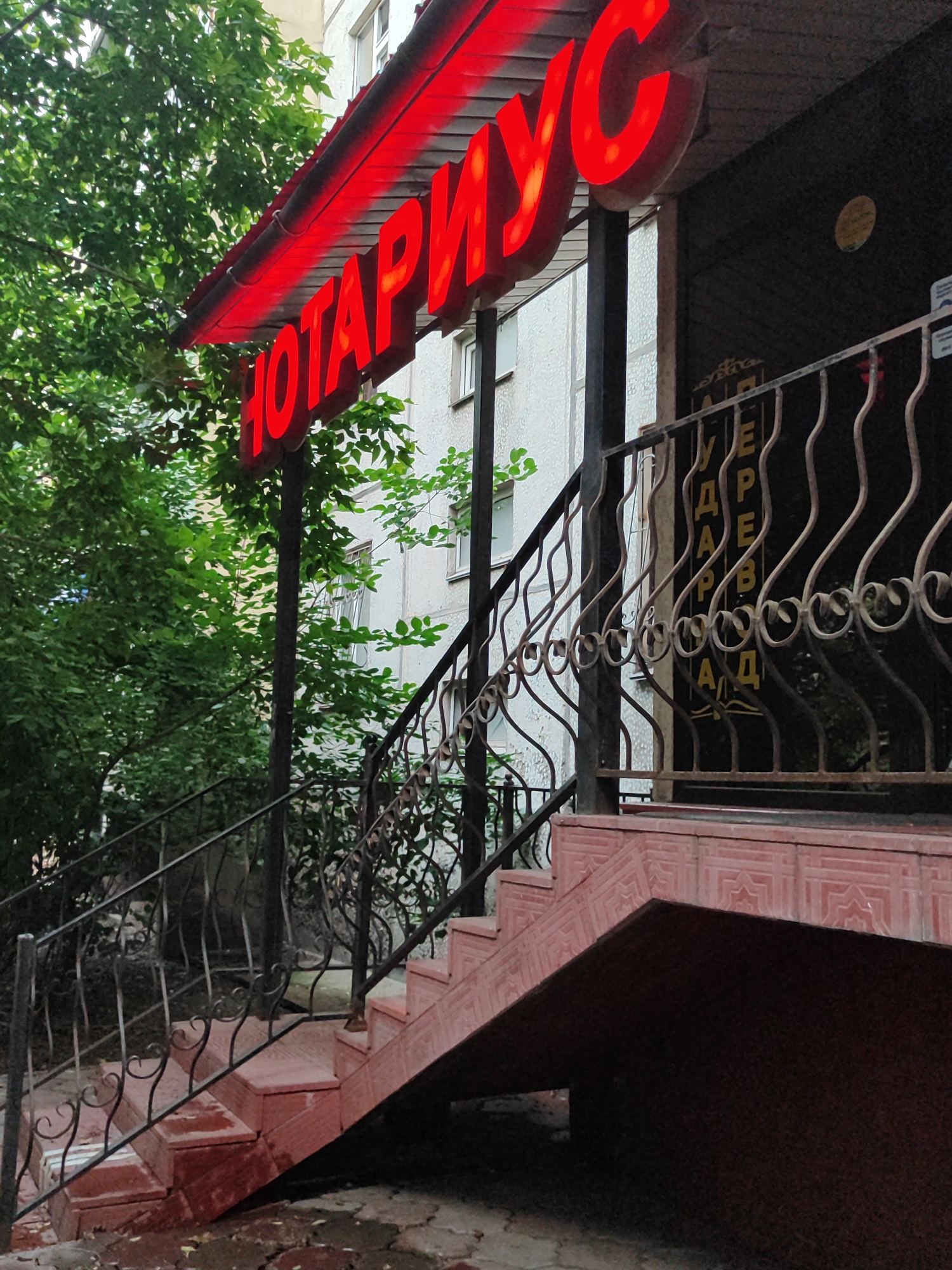 The media file depicts the exterior of a building with a red neon sign that reads "НОТАРИУС" (Notary). The sign is illuminated and positioned above a staircase. The staircase is made of red material and has black metal railings. Lush green trees surround the entrance, suggesting an outdoor setting. The building itself appears to be a multi-story structure with windows visible. On the right side of the image, behind the railing, there is a dark facade with some lettering on a lighter background, which appears to be "УДАР" or similar Russian letters. The time of day appears to be daytime, possibly late afternoon or early evening, given the lighting and the shadows. The weather seems to be mild, as indicated by the foliage. The overall impression is of a functional building entrance, likely a notary's office, in an urban environment in Almaty, Kazakhstan. There is no visible activity or people in the frame.