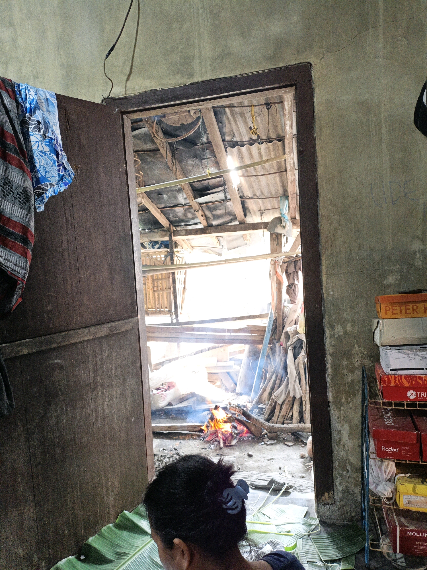 The image depicts an interior scene looking through an open doorway into an outdoor or semi-enclosed area where a fire is burning. The room in the foreground has a rough, textured wall. A wooden door, partially ajar on the left, has clothing draped over it, including a patterned blue and white fabric with red and grey stripes. A person's head and shoulders are visible in the lower foreground, facing away from the camera and towards the doorway. They have dark hair pulled back with a light blue clip and are positioned near a spread of large green leaves, possibly banana leaves.
Through the doorway, the scene is brighter and appears more rustic. The fire is a prominent feature, with visible flames and smoke rising. Logs and branches are scattered around the fire pit. The area beyond the fire seems to be constructed with wood and corrugated roofing material, suggesting a simple structure. Various objects, including piles of wood and what appear to be sacks or bags, are present. Sunlight streams into this area from the right.
On the right side of the foreground room, a multi-tiered shelf unit holds several stacked boxes, some of which have visible labels, including "PETER K," "Fladeo," and "MOLLIN." The overall environment suggests a humble dwelling or workspace in a rural or less developed setting, consistent with a location in Bunut, Indonesia.