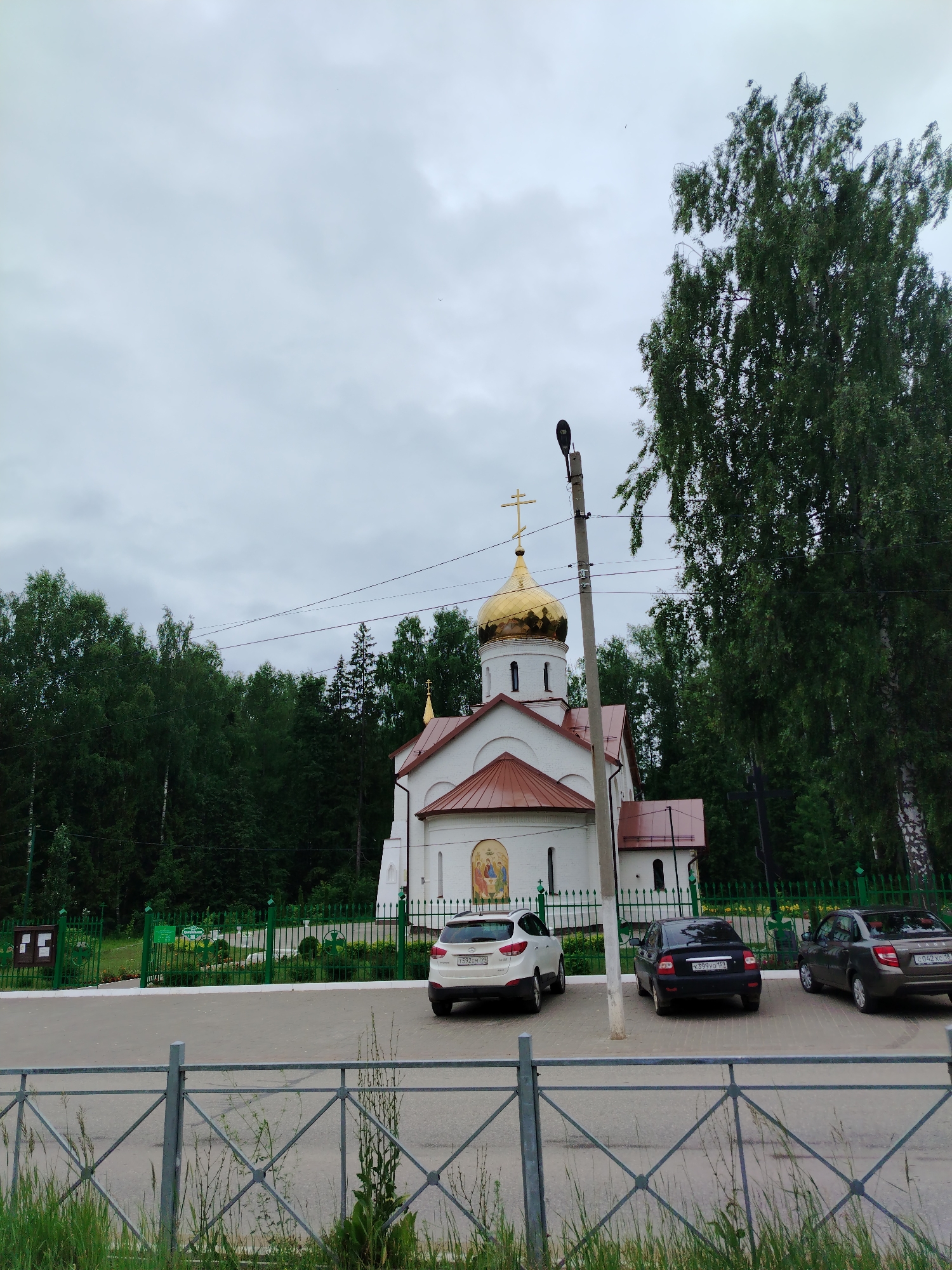 A white church with a golden dome and a red roof is visible in Noviy, Russia. The church has a fresco above its entrance and a cross on top of the dome. Several cars are parked in front of the church, including a white SUV, a black sedan, and a gray sedan. The church is surrounded by a green fence and a line of trees. The sky is overcast and the weather appears to be cloudy.