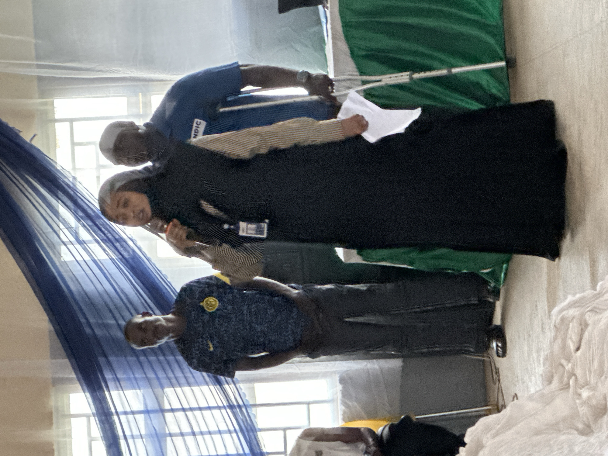 The image, rotated for correct viewing, captures three individuals standing indoors in a brightly lit room. In the center, a woman wearing a black abaya and hijab smiles faintly, appearing to hold a phone or small object to her ear. An ID badge hangs from her hip. Behind her, a man in a blue polo shirt holds a white piece of paper. The acronym "NDIC" is clearly visible on the front of his shirt. To the woman's left and slightly in front, another man in a dark blue patterned shirt, sporting a yellow circular logo on his chest, observes the scene.
The setting appears functional, with large windows in the background allowing bright daylight to stream in, indicating it is daytime. A sheer blue fabric, likely a curtain or mosquito net, is visible on the left side of the frame. On the right, a dark green fabric or surface is present, with a silver crutch or walking stick leaning against it. A pile of white fabric or sheets can be seen in the bottom right corner. The presence of the "NDIC" branding suggests a connection to the Nigeria Deposit Insurance Corporation, aligning with the Unguwan Dosa, Nigeria, location context. The individuals seem engaged in an interaction, possibly an informal meeting or an event.