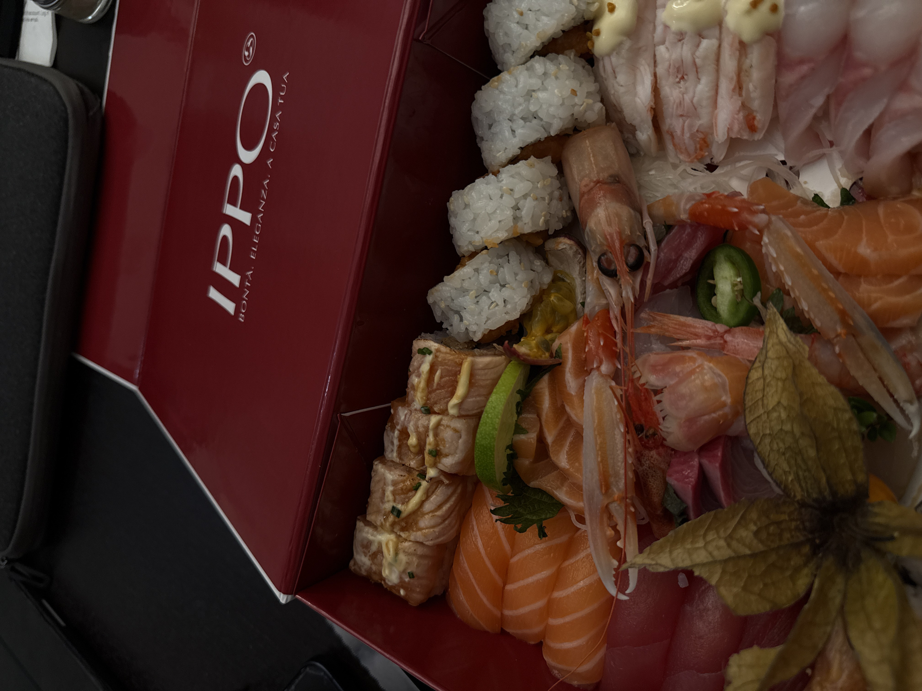 A red box with "IPPO. BONTÀ. ELEGANZA. A CASA TUA" printed on the side is filled with assorted sushi and sashimi. There are California rolls, rolls drizzled with sauce, salmon sashimi, white fish sashimi, and various types of seafood, including shrimp and scampi. Garnishes include a slice of lime, jalapeño, and a dried fruit with papery leaves.