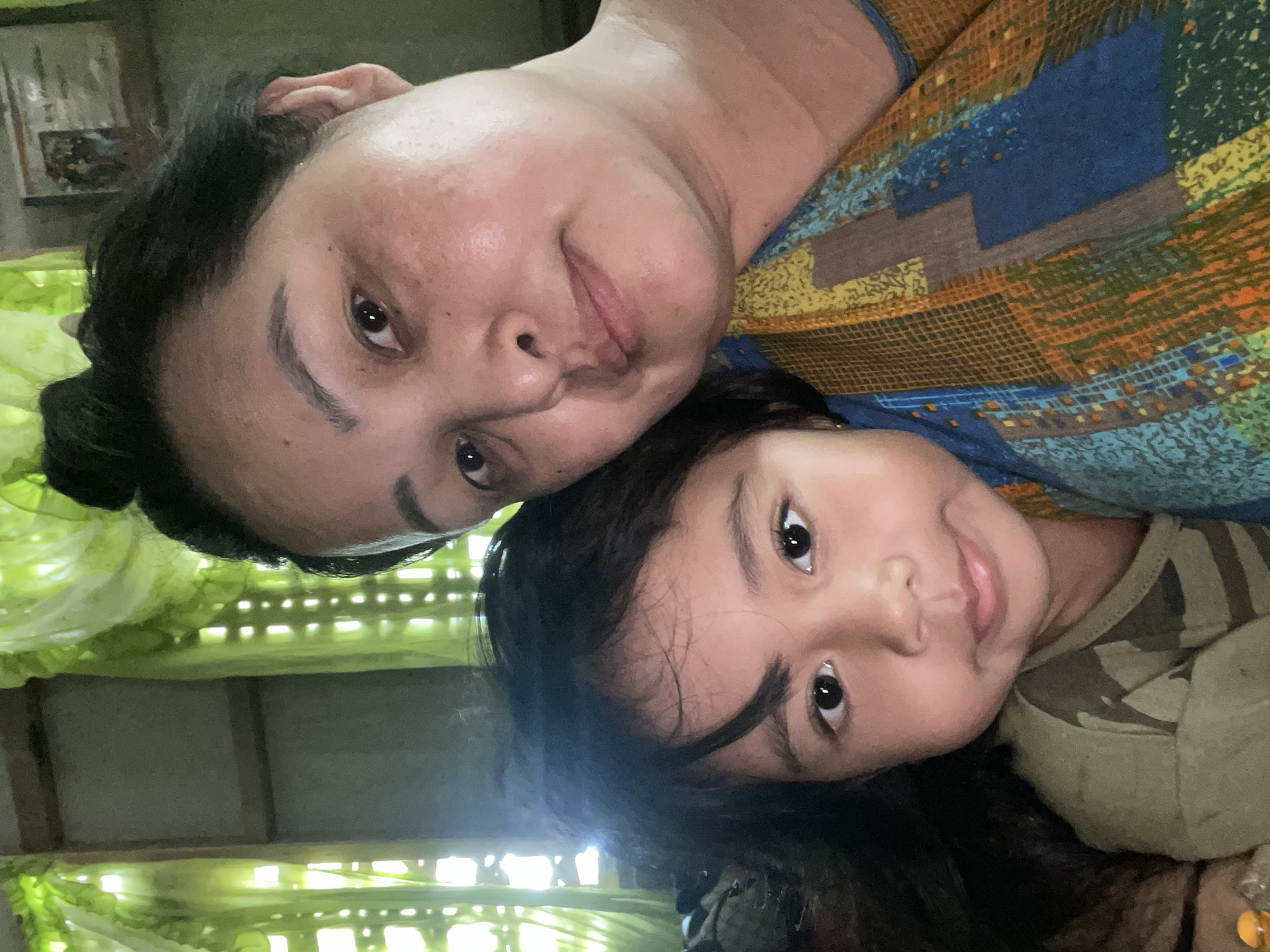 The image depicts a selfie taken indoors, featuring an adult and a child, likely a parent and child. The adult, positioned in the upper portion of the frame, has dark hair pulled back, and is wearing a patterned top with blue, yellow, and brown hues. The child, located beneath the adult, has dark hair and is wearing a green camo-patterned shirt. The background suggests a room with green curtains or foliage visible through window bars or a patterned screen. There is no explicit evidence within the image to confirm the location is in Hijo, Philippines.
