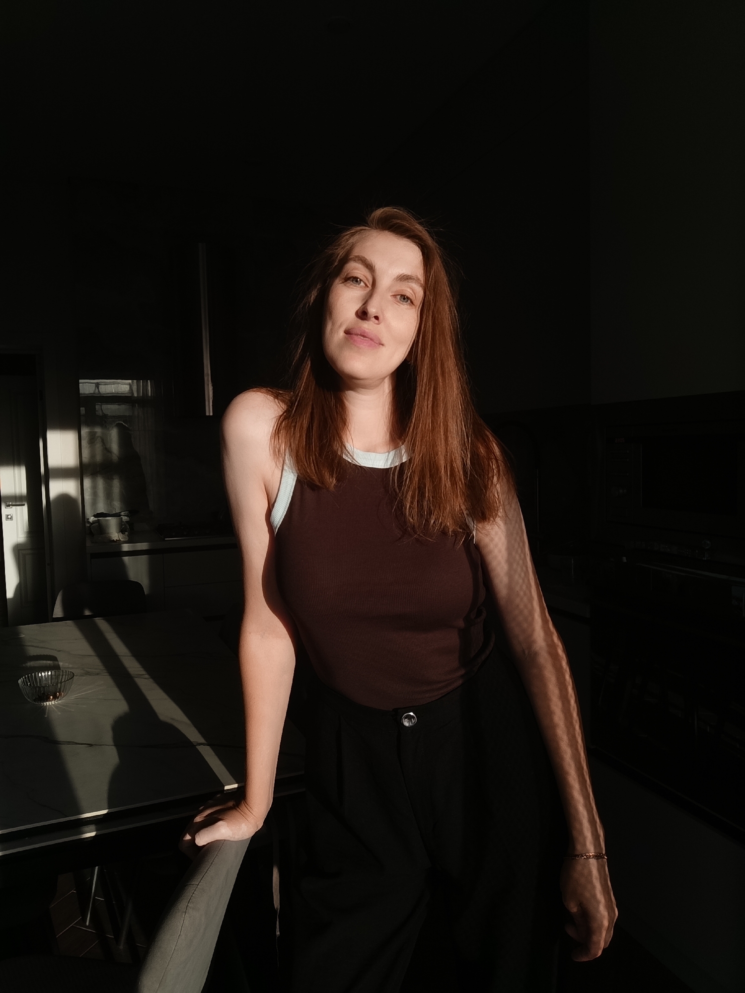 The image depicts a young Caucasian woman with reddish-brown hair and light-colored eyes, indoors in what appears to be a kitchen. She is wearing a brown tank top with a light blue trim and black pants. Sunlight streams in from the left, casting strong shadows across the scene, including a checkered pattern on her arm and shoulder. She is leaning on a chair next to a table. The background is dark, showing kitchen cabinetry and appliances. A window with vertical blinds is visible on the left, allowing the sunlight to enter. The location is identified as Chaikovsky, Russia.