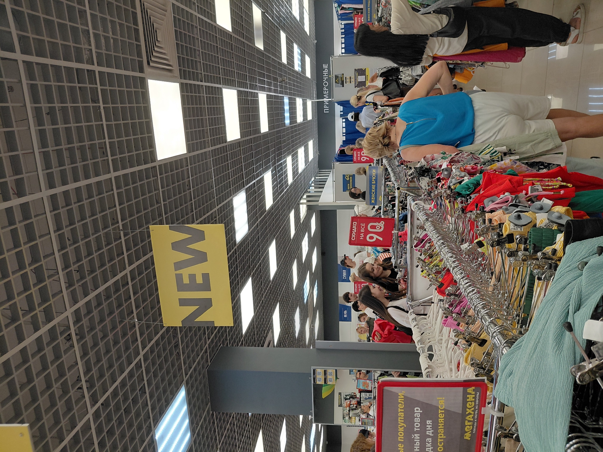 In a brightly lit retail store, likely a clothing department, a yellow sign with the word "NEW" in bold, gray letters hangs from the ceiling. The ceiling itself is a grid of white acoustic tiles with recessed fluorescent lighting panels. Racks of clothing are organized throughout the store, with a variety of colors and styles visible. In the foreground, a rack of pastel-colored clothing, including a light blue textured fabric, is prominent. Several shoppers are browsing the merchandise, with one woman in a blue top and white shorts visible from the back. Another woman, wearing a black top, is standing further down an aisle, looking at clothing. Signs indicating sales and departments, such as "СКИДКА НА ВСЁ 90%" (Discount on everything 90%) and "ПРИМЕРОЧНЫЕ" (Fitting rooms), are present. The overall impression is of a busy and well-stocked retail environment.