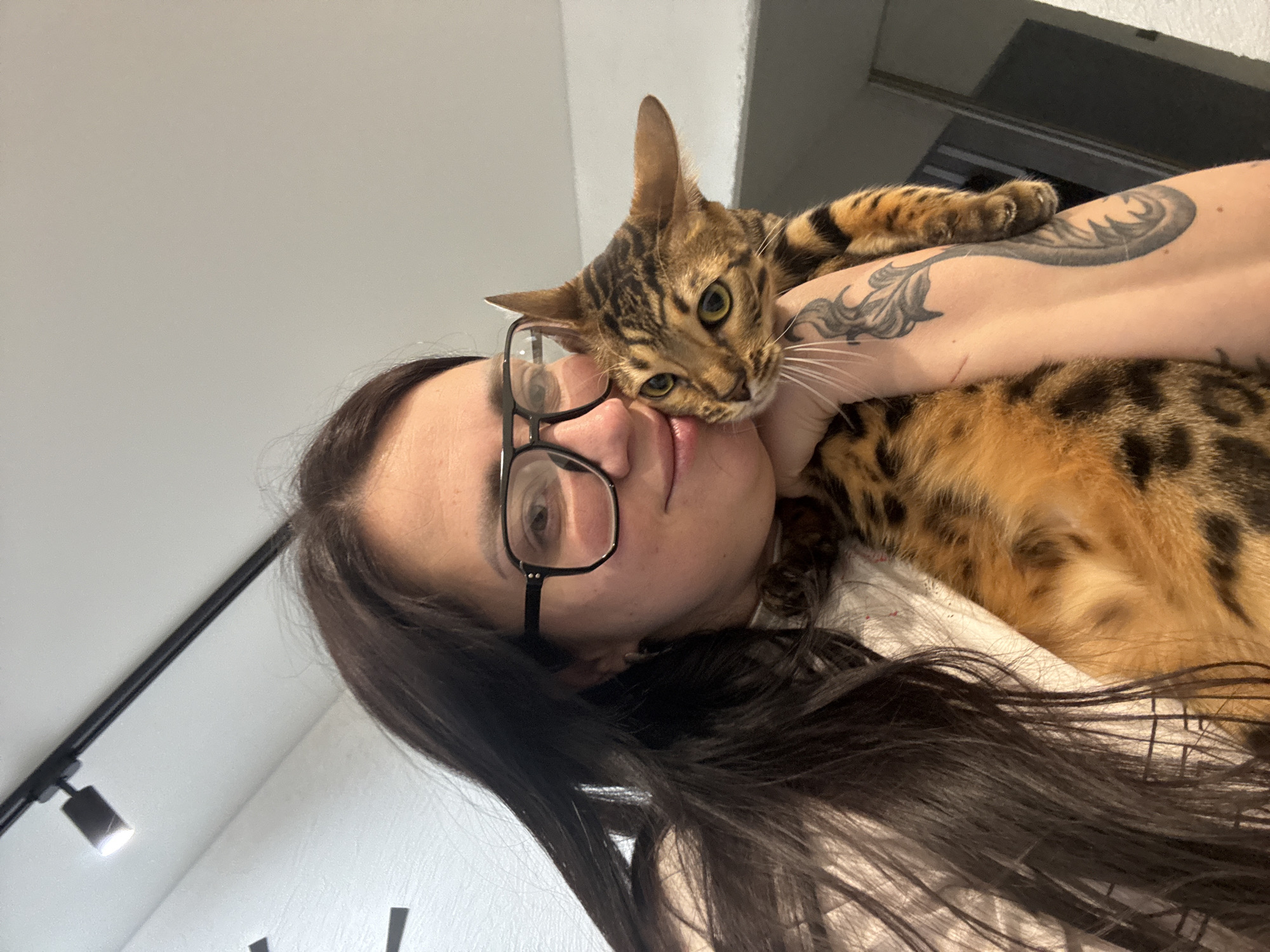 A woman with dark hair and glasses holds a Bengal cat against her face, indoor. The woman is wearing a light-colored shirt. The cat is on the woman's arm, which has a tattoo on it. The background includes a white wall, a track lighting fixture, and a glimpse of an open doorway. The location in the city of Unknown, Unknown, is not possible to infer from the image.