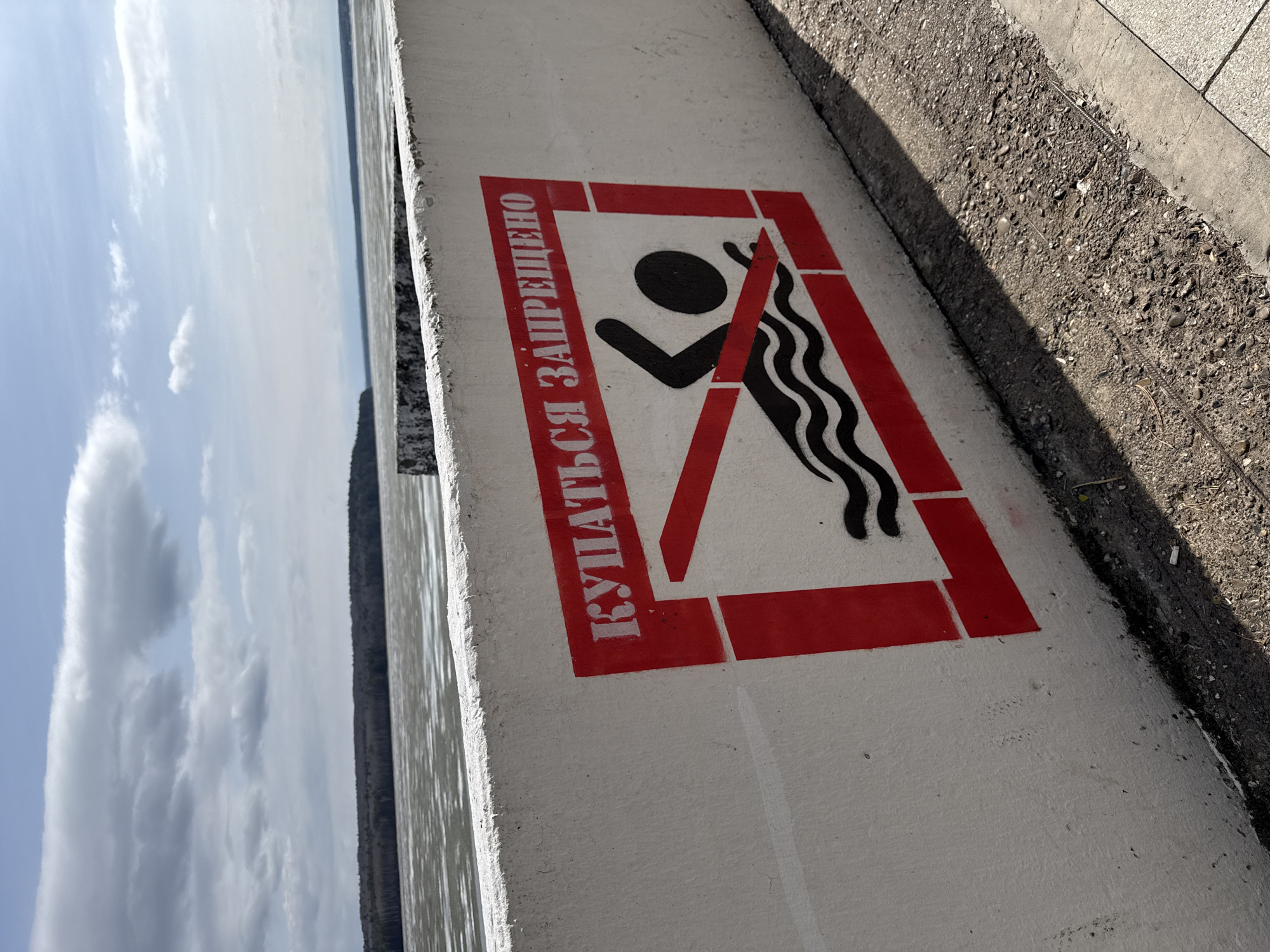 The image shows a concrete wall along a body of water. On the wall is a red and white sign indicating that swimming is prohibited. The sign depicts a stick figure swimming with wavy lines representing water, with a red line crossing over the figure. The text on the sign is in Russian, translating to "Swimming is Prohibited." The location is near Chaikovsky, Russia. Adjacent to the wall is a section of gravel and a paved walkway. The sky is visible in the background.