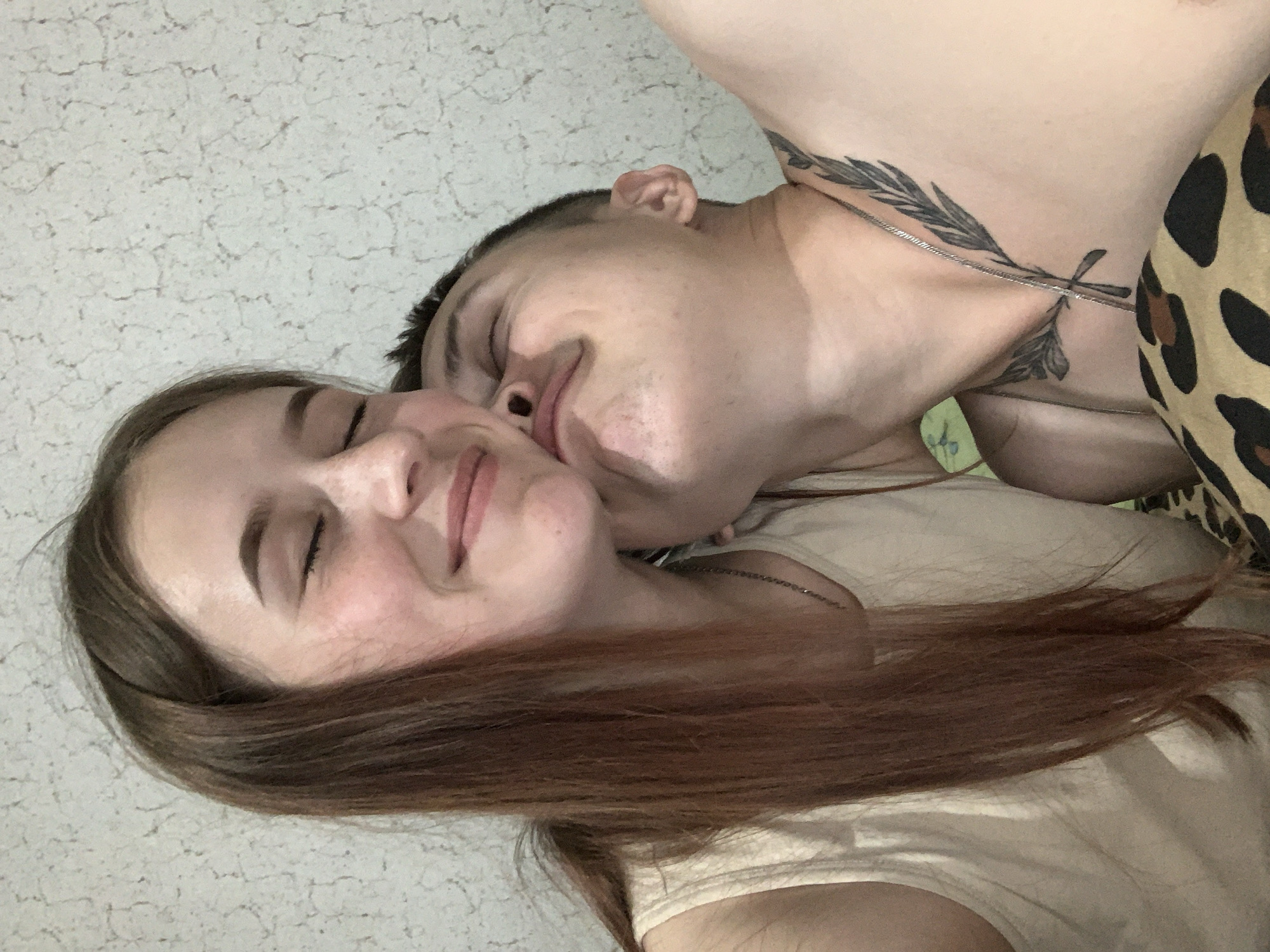 A horizontal photograph shows two individuals in a close embrace. The person on the left, presumably a female, has light skin, closed eyes, and light brown hair. The person on the right, presumably a male, has tan skin, closed eyes, short dark hair, and a tattoo on the neck. The male is kissing the female on the cheek. Both are wearing light-colored clothing. In the background is a wall with a light texture. The photo's location is unknown and cannot be confirmed to be Фоки, Russia.