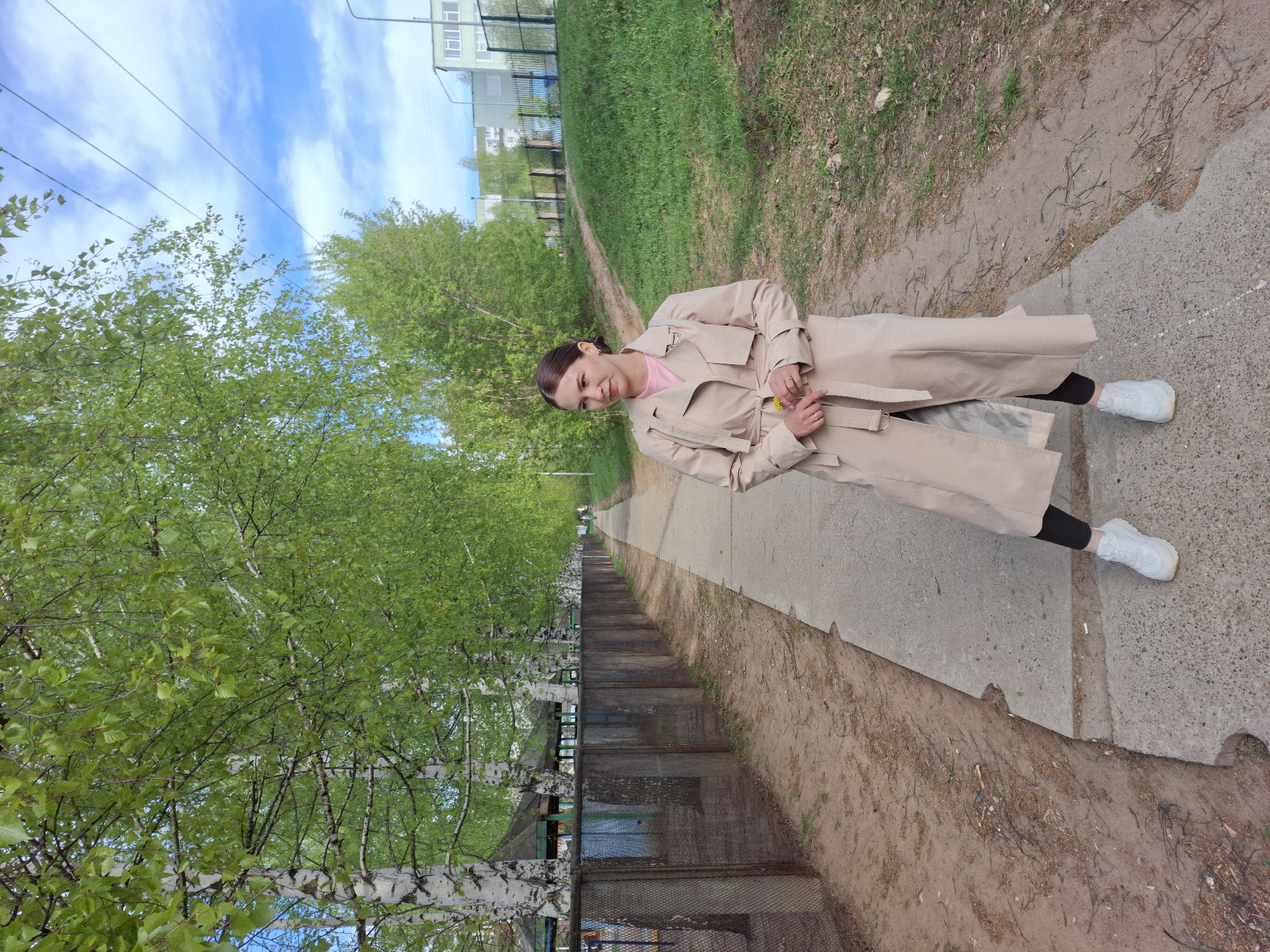 The photograph depicts a woman standing on a concrete pathway in what appears to be a park or green area. The woman is wearing a light-colored trench coat over what seems to be a pink top and black leggings, and white sneakers. She is holding a small yellow flower.
To her left, there is a row of birch trees. Below the trees is a metal fence and a small green roofed structure, and below that is a series of steps. To her right is a dirt or grassy bank.
The background includes more greenery and buildings. The sky is blue with some clouds.
Based on the location metadata, the image was taken in the city of Chaikovsky, Perm Krai, Russia.