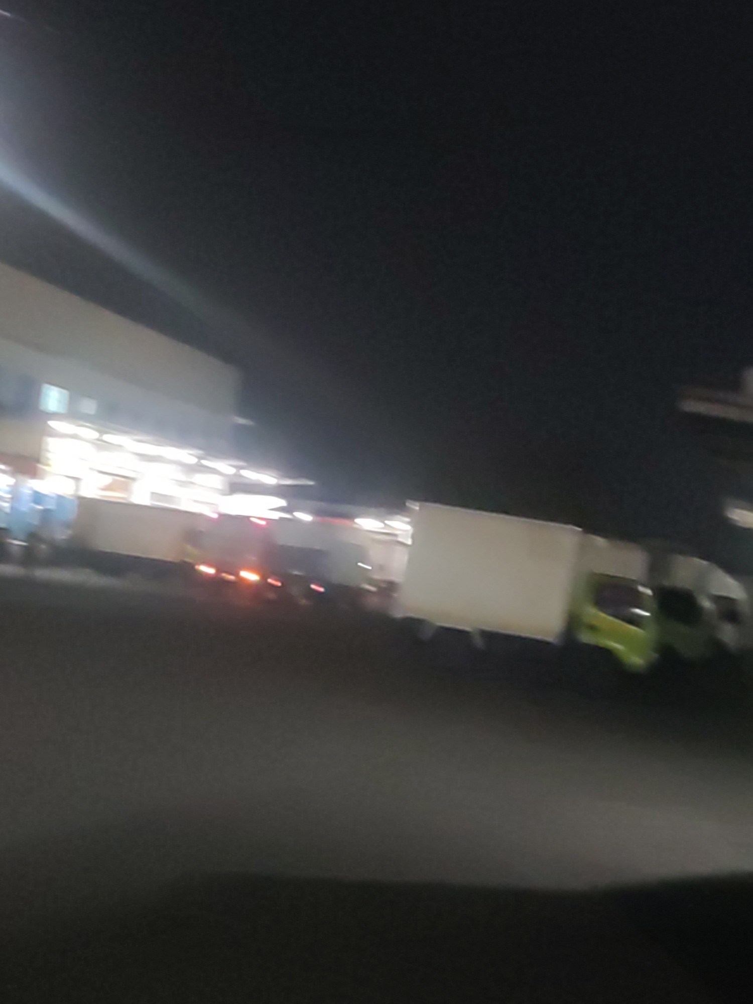The image depicts a dimly lit scene at night, possibly in a commercial or industrial area of Trihanggo, Indonesia. A building with illuminated windows or lights is visible on the left, suggesting it might be a warehouse or a commercial establishment. In front of the building, there are several parked vehicles. One vehicle, appearing to be a truck, is white with visible red brake lights. Another vehicle, a lighter colored truck with a green cabin, is also present. The ground is paved, and the overall impression is one of a logistics or transportation hub operating during nighttime hours. The image is blurry, making it difficult to discern specific details about the vehicles or the surrounding environment.