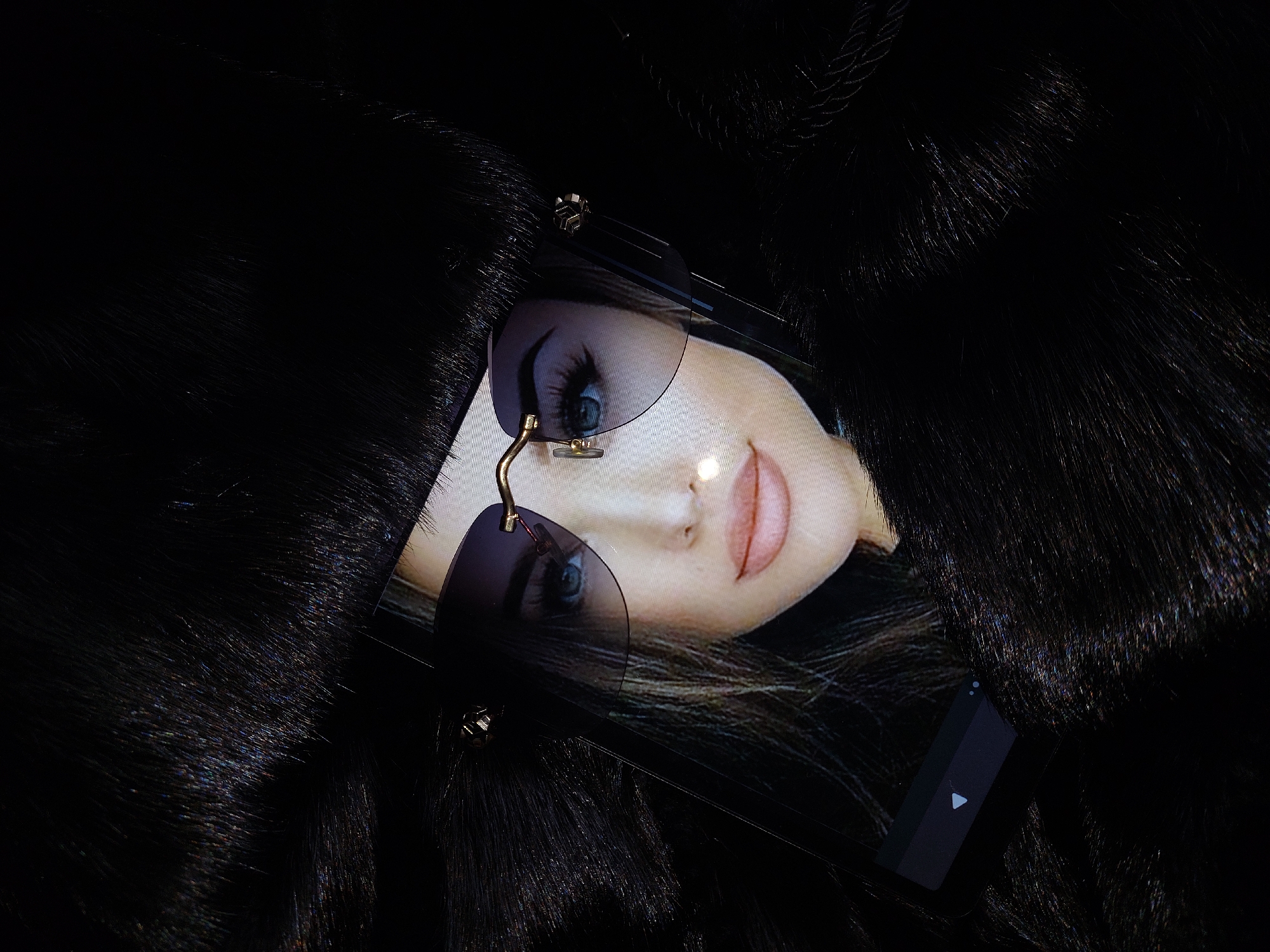 A cell phone displays a woman's face wearing frameless sunglasses. The phone is partially obscured by dark, fluffy material, possibly fur. The background is dark. The image does not provide enough information to determine a location in Chaikovsky, Russia, or any other specific details about people, actions, or interactions.