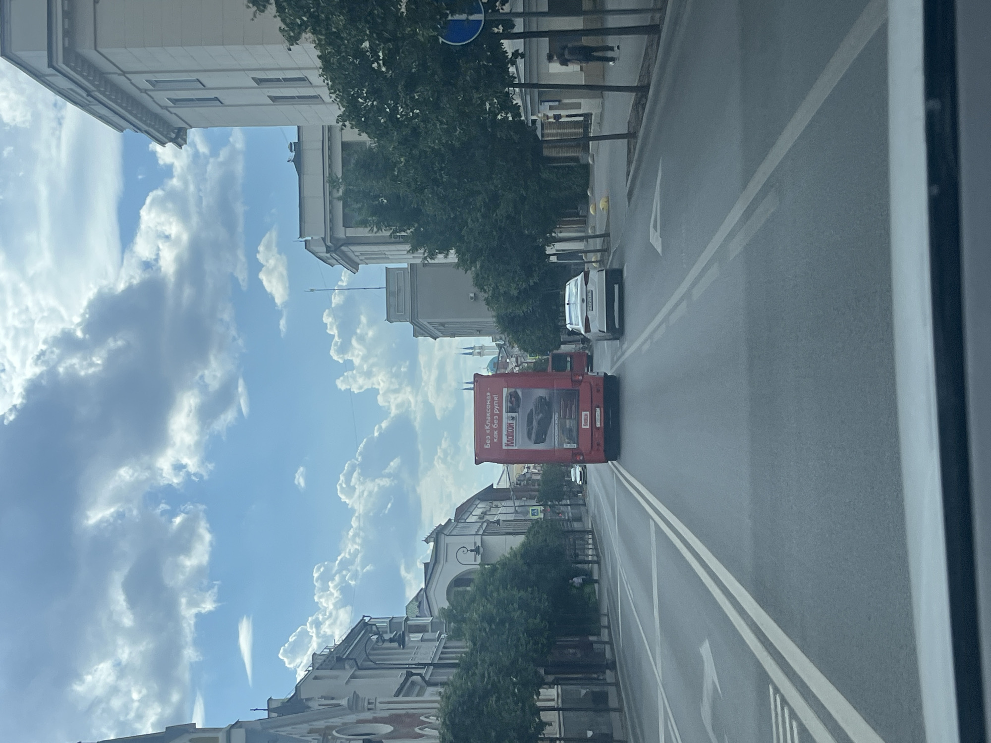 The image depicts a street scene in Kazan, Russia, on a partly cloudy day. The perspective is from a vehicle, looking forward down a multi-lane asphalt road. On the right side of the road, there is a sidewalk lined with trees and buildings. A white sedan is traveling in the same direction as the viewer, and further ahead, a red truck with advertising on its back is also visible. The truck has text and images, including a car advertisement. On the left side of the road, there are more trees and buildings, some of which appear to be older architecture. A few pedestrians are present on the sidewalks. The sky is filled with dynamic clouds, suggesting fair weather. The presence of Cyrillic script on the red truck, along with the distinctive architecture of the buildings and the general urban environment, indicates the location is in Russia, likely Kazan, given the prompt.