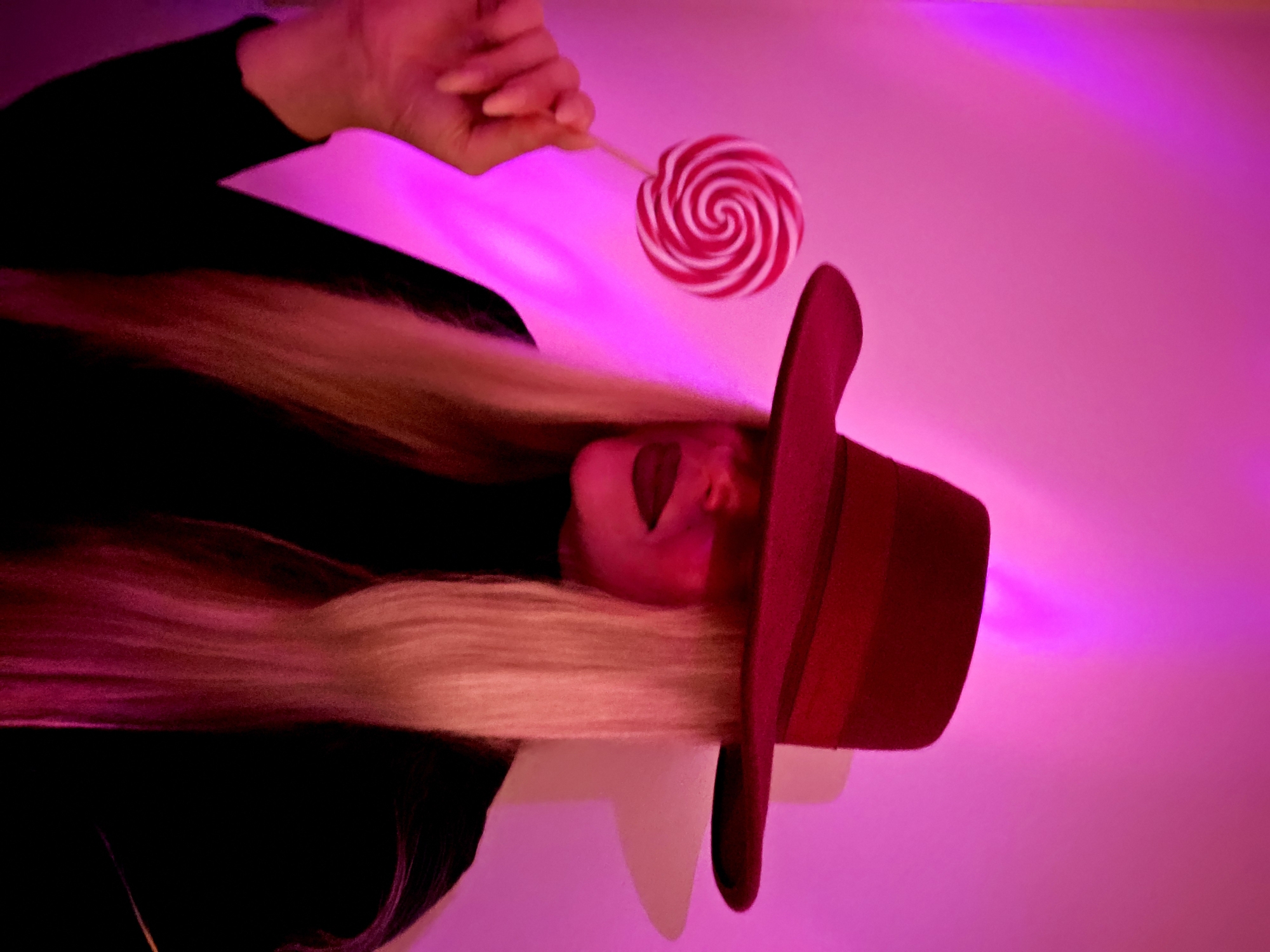 The image depicts a person, likely female, with long blonde hair, wearing a black top and a wide-brimmed, dark red hat. The original image is rotated 90 degrees clockwise. The person's face is partially obscured by the hat and hair, but their mouth is open as if interacting with a red and white swirl lollipop. A hand is holding the lollipop on a stick, presenting it to the mouth.
The entire scene is illuminated by a strong, artificial fuchsia or magenta light, casting a vibrant glow across the background and partially on the subject's hair and hat. The background appears to be a plain wall or backdrop uniformly colored in this bright pink hue. The lighting creates a dramatic and stylized effect.
No specific location cues, time of day, or weather elements are discernible, suggesting an indoor or studio setting. There is no visible text in the image. The overall impression is one of playful indulgence and striking visual aesthetics due to the bold color scheme and dynamic pose.