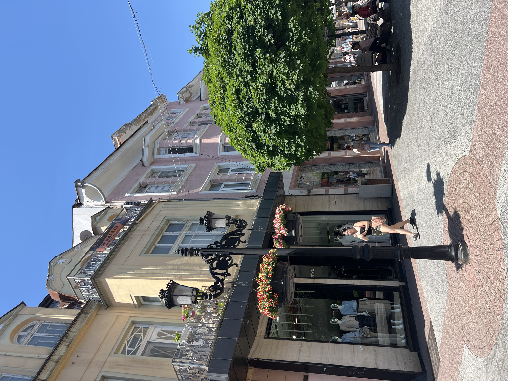 A street scene in Plovdiv, Bulgaria. The image is taken at street level, focusing on the architecture and pedestrian activity. The buildings are a mix of light yellow and pink and white colors, with traditional European-style facades and ornate balconies. There is a large tree in the upper right corner of the image.
A woman is standing on the sidewalk in front of a storefront, looking at her phone. A dark-colored lamppost with hanging flower baskets stands nearby. The sidewalk is paved with red and grey bricks in a circular pattern. In the background, people are sitting at tables under the shade of trees. The sky is clear and blue.