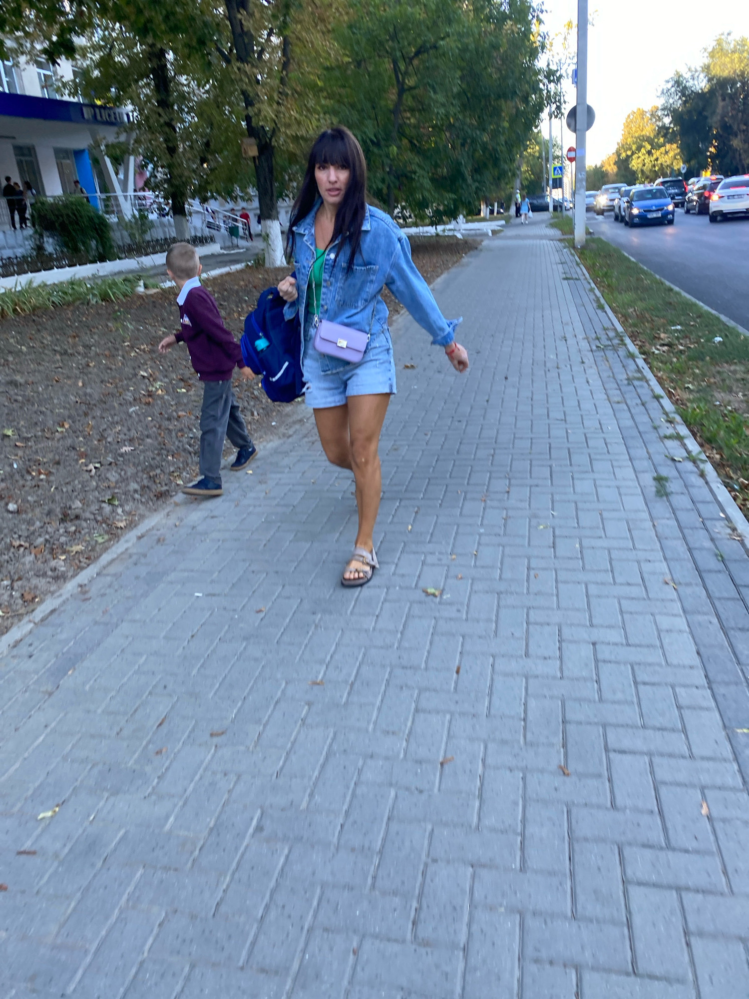 A woman and a young boy are walking on a sidewalk next to a road with traffic in Chișinău, Moldova. The woman, who appears to be in her 30s, is wearing a denim jacket, green top, and denim shorts. She is carrying a blue backpack and a small purple purse. The boy, who is younger than 10 years old, is wearing a maroon sweater and dark pants. They are walking away from a building that appears to be a school, with "LICEUL" visible on the facade. The road has several cars driving on it, and the sidewalk is paved with grey bricks. Trees line the street, and the overall setting suggests an urban environment in the autumn, with some fallen leaves on the ground. The sun is shining, casting long shadows.