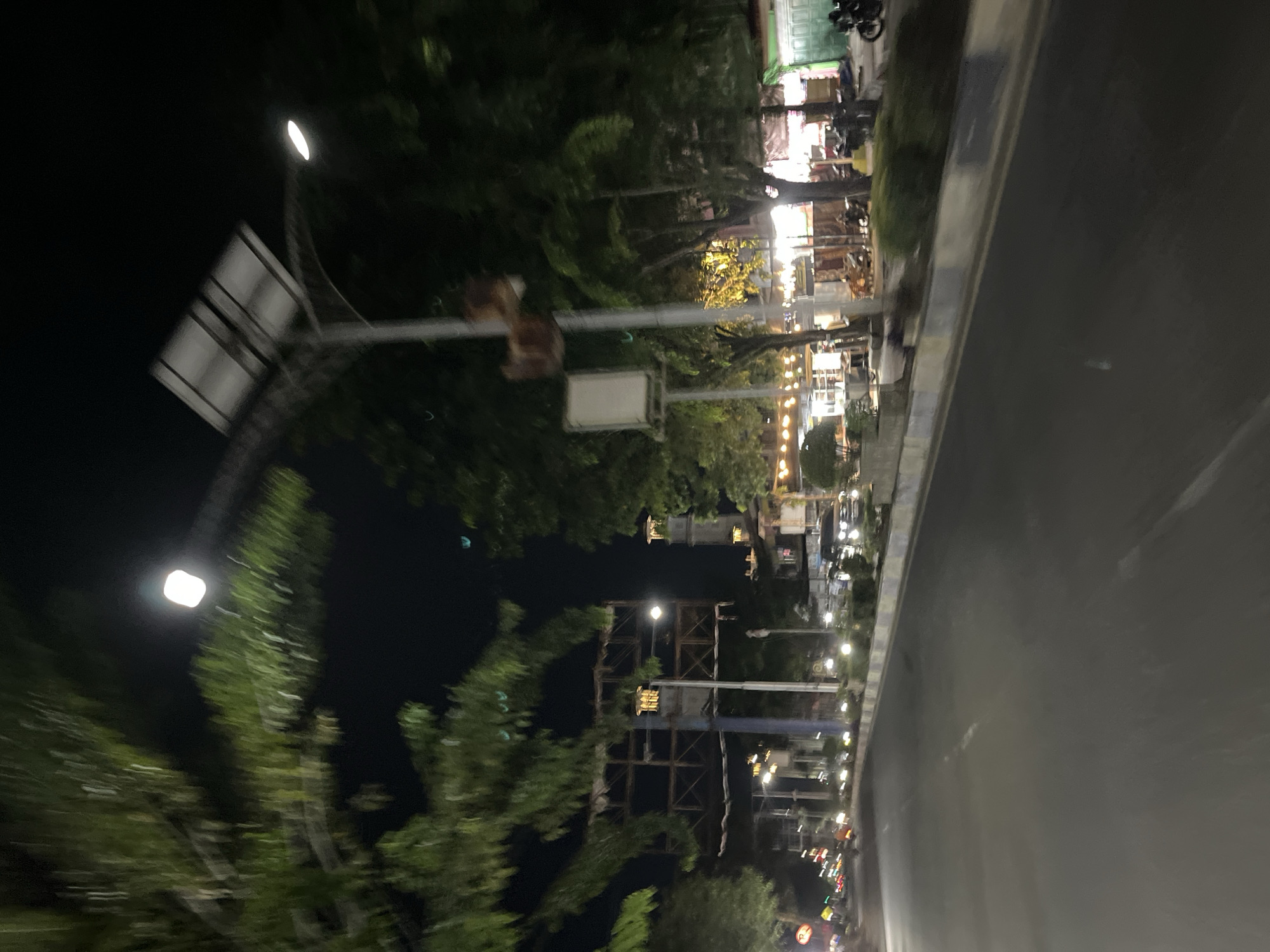 The image shows a street scene at night in Langsa, Indonesia. A road occupies the foreground, with vehicles and streetlights visible on the right side. Trees and some structures line the left side of the road. A lamppost with a solar panel is prominent in the upper right quadrant of the image. The overall impression is of a well-lit urban street at night.