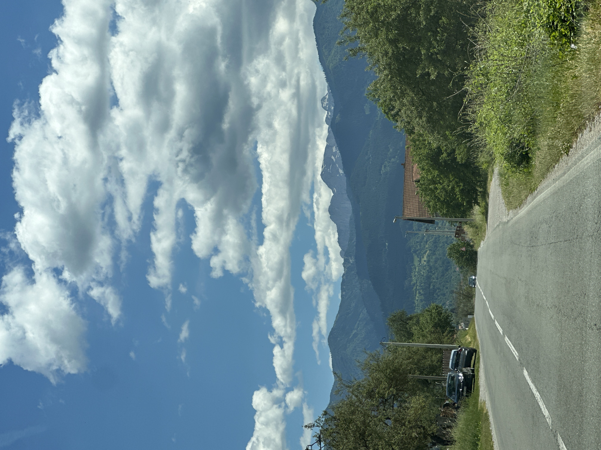 A paved road curves gently to the right, leading the viewer's eye towards a vast mountain range under a partly cloudy sky. Two cars, a dark sedan and a lighter colored SUV, are parked on the left side of the road near the foreground. Green trees and lush grass line the sides of the road. In the middle ground, a house with a red-tiled roof is nestled amongst dense trees. The mountain range in the background is covered in varying shades of green, with some snow-capped peaks visible in the distance. The sky is a vibrant blue with scattered white and gray clouds. The scene depicts a rural or mountainous area, possibly near Murino, Montenegro, during daylight hours.