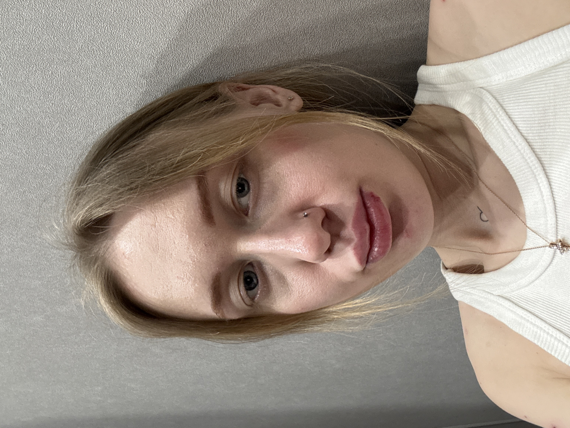 A young, light-skinned female with blonde hair is depicted in a close-up, head-and-shoulder shot. She has blue eyes and a small silver stud piercing her left nostril. Her lower lip appears swollen, exhibiting a prominent red lesion or abrasion on its right side, with minor associated redness on the chin below. She is wearing a white ribbed tank top. A small black, crescent-shaped tattoo is visible on the left side of her neck, and a delicate gold-colored chain with a small crucifix pendant hangs around her neck. Small red marks or scabs are present on both of her shoulders/upper arms. The subject is positioned against a plain, textured gray wall, indicating an indoor environment. She is looking directly towards the viewer.