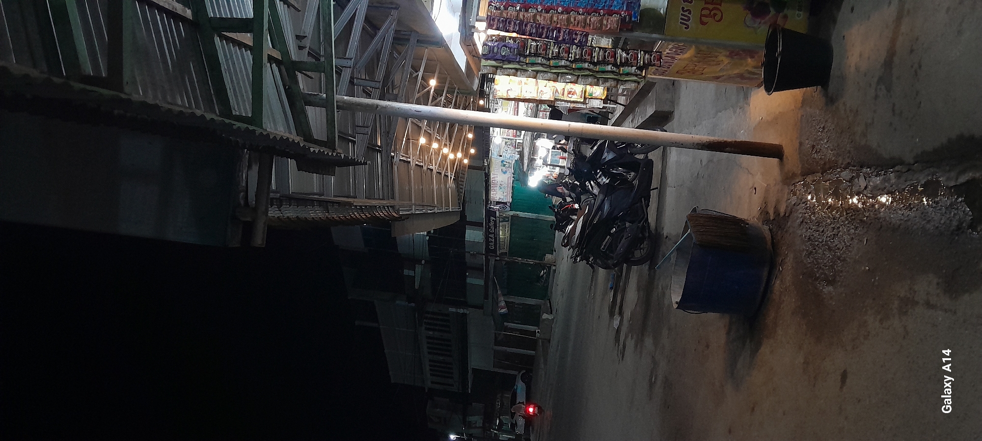 This is a nighttime, eye-level photograph taken on a street in Keudegeudong, Indonesia. The scene is dimly lit by artificial lights, with a row of shops on the right side of the frame and buildings in the background. In the foreground, a dark blue plastic bucket with a broom inside sits on the wet asphalt. A small pool of water reflects the lights from the shops.
The shops appear to be open, with goods displayed on shelves and racks. There are several motorcycles parked in front of the shops, and one motorcycle with its taillight illuminated is leaving the frame on the left. A person is visible behind the parked motorcycles, possibly a vendor or customer. The architecture of the shops features corrugated metal roofing supported by metal posts.
The overall impression is of a typical street scene in a bustling Indonesian town at night, with the presence of commerce and transportation evident. The wet ground suggests recent rain. The "Galaxy A14" watermark is present in the bottom left corner of the image.