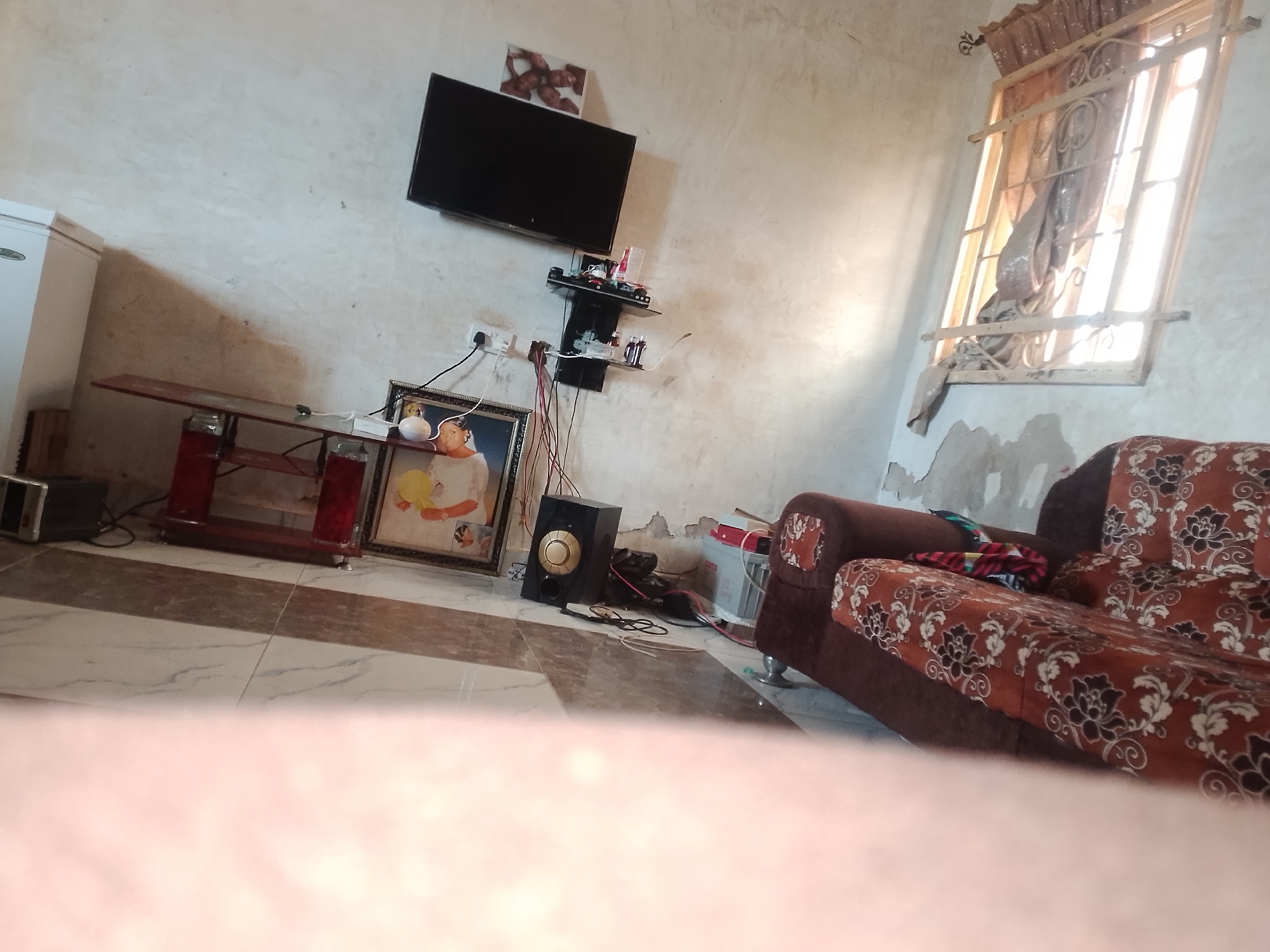 This media file depicts an indoor scene, likely a living room, in Jalingo, Nigeria. The room has a somewhat unkempt appearance with cracked and textured walls.
Dominating the view is a brown armchair with a floral pattern in shades of white and black. A red and black patterned throw is draped over the armrest. To the left of the armchair, on the floor, is a black speaker. Further to the left, a framed picture hangs on the wall. The picture appears to be a portrait of a woman in a white dress and a yellow sash, with another figure partially visible.
Mounted on the wall above the picture is a flat-screen television. Below the television are two small shelves holding various items, including what appear to be bottles and electronic components. A white refrigerator is visible on the far left of the image.
A large window with metal bars and a curtain is situated on the right side of the frame, allowing bright daylight to enter the room. The floor is tiled with a pattern of light and dark brown squares. The overall impression is of a lived-in space, possibly a residential setting, during daylight hours. There is no clear indication of a specific activity or event taking place.