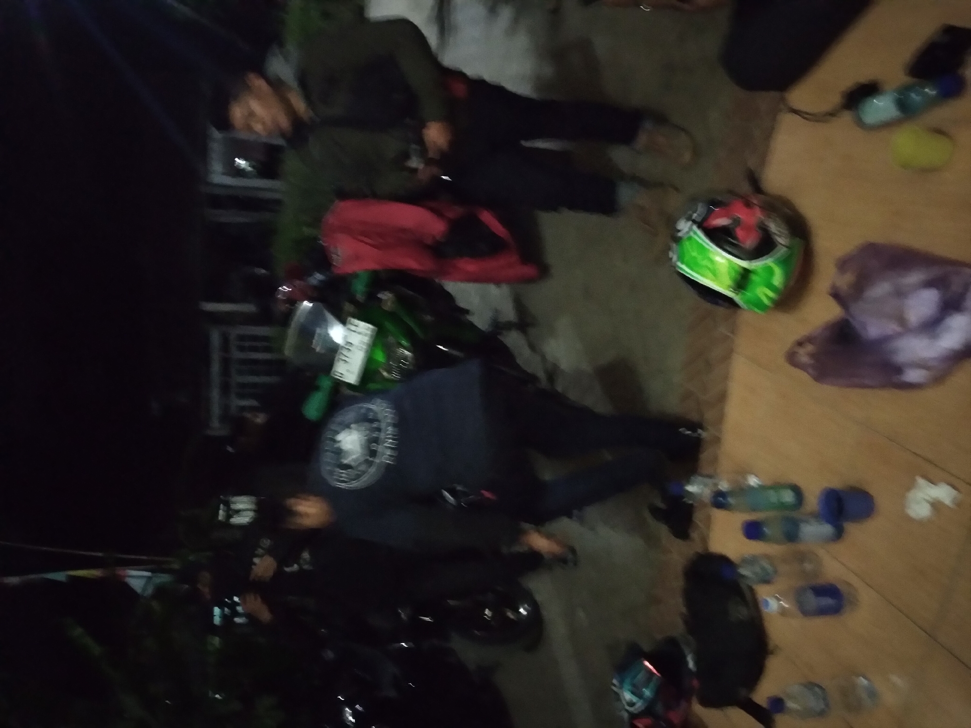 This image depicts a nocturnal scene, likely in the city of Pandak, Indonesia, with a group of individuals gathered around motorcycles. The lighting suggests it is nighttime, with artificial lights illuminating the area.
In the foreground, several water bottles are scattered on a wooden surface, along with a black bag and a vibrant green and red motorcycle helmet. A few other objects, including a purple bundle and a yellow object, are also visible.
Behind the foreground objects, three individuals are present. One person, wearing a dark jacket and pants, stands near a motorcycle, looking towards the right of the frame. Another individual, in a dark blue long-sleeved shirt with a circular logo on the back, is bending down. A third person, wearing a dark jacket, stands to the right.
Several motorcycles are present, including a prominent green one with a license plate. In the background, structures that appear to be buildings or shelters are visible, along with foliage and what might be flagpoles. The overall atmosphere suggests a casual gathering or break during a nighttime activity, possibly related to motorcycling.