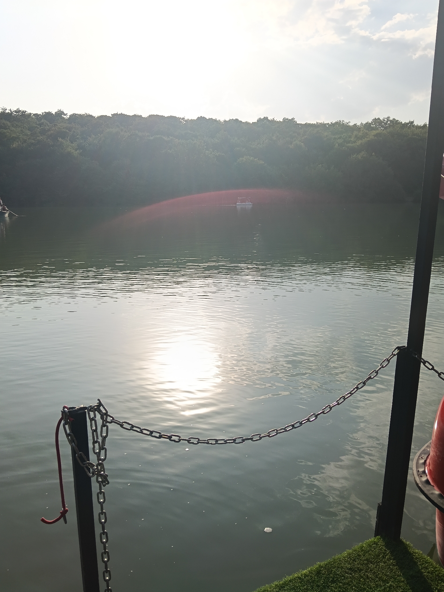 The image depicts a serene lake scene on a sunny day. In the foreground, a black metal post is secured with chains, and a red rope is tied to it. The post is situated on a patch of artificial green turf. The lake stretches out into the mid-ground, with gentle ripples on its surface reflecting the sunlight. In the distance, a small white paddleboat can be seen on the water. The background is dominated by a dense line of trees, forming a natural border to the lake. The sky above is bright with scattered clouds, indicating fair weather. The overall impression is one of tranquility and natural beauty, likely a recreational area in or near Köhnə Xudat, Azerbaijan.