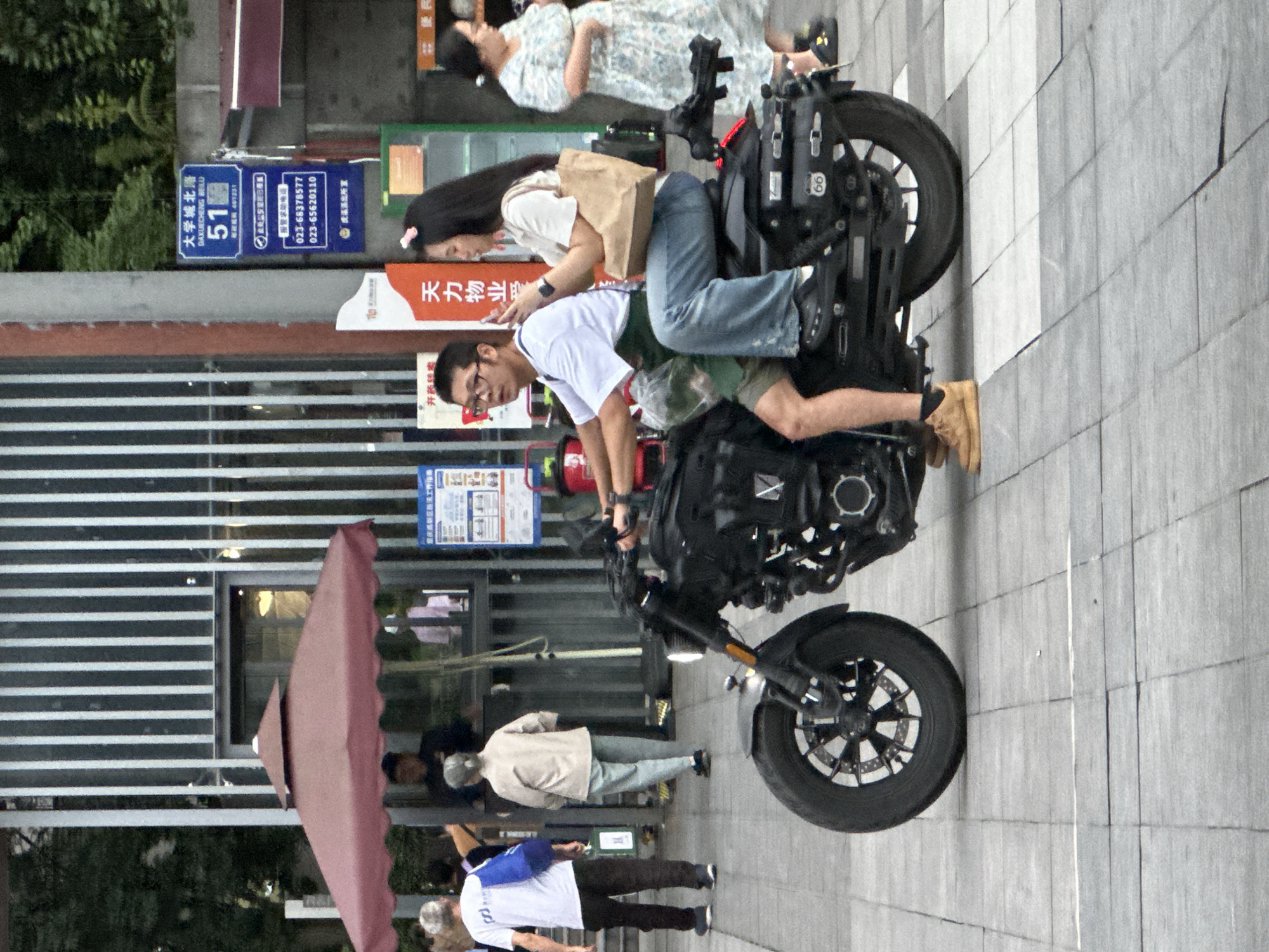 The image depicts a street scene in Chongqing High-tech Zone, China. The main subjects are a young couple on a motorcycle. The man is driving, wearing a white t-shirt and glasses, while the woman is a passenger behind him, wearing a white t-shirt and blue jeans. She is holding a brown paper bag and her phone. The motorcycle is black and appears to be a custom model.
In the background, several other people are visible, some walking and others on what appear to be scooters or motorcycles. A red umbrella is also prominently displayed on the left side of the frame.
Several signs are visible in the image. One blue sign reads "大学城北路 DAXUECHENG BEILU 51". Another sign displays phone numbers: "023-68378577" and "023-65620110". The text "天力物业" and "开药转卖" are also present on signs.
The scene appears to be set during the daytime, likely in an urban environment with a mix of commercial and residential buildings. The lighting suggests it might be overcast or late afternoon. The overall atmosphere seems casual, with people going about their day. The people on the motorcycle do not show any strong emotions in their poses.