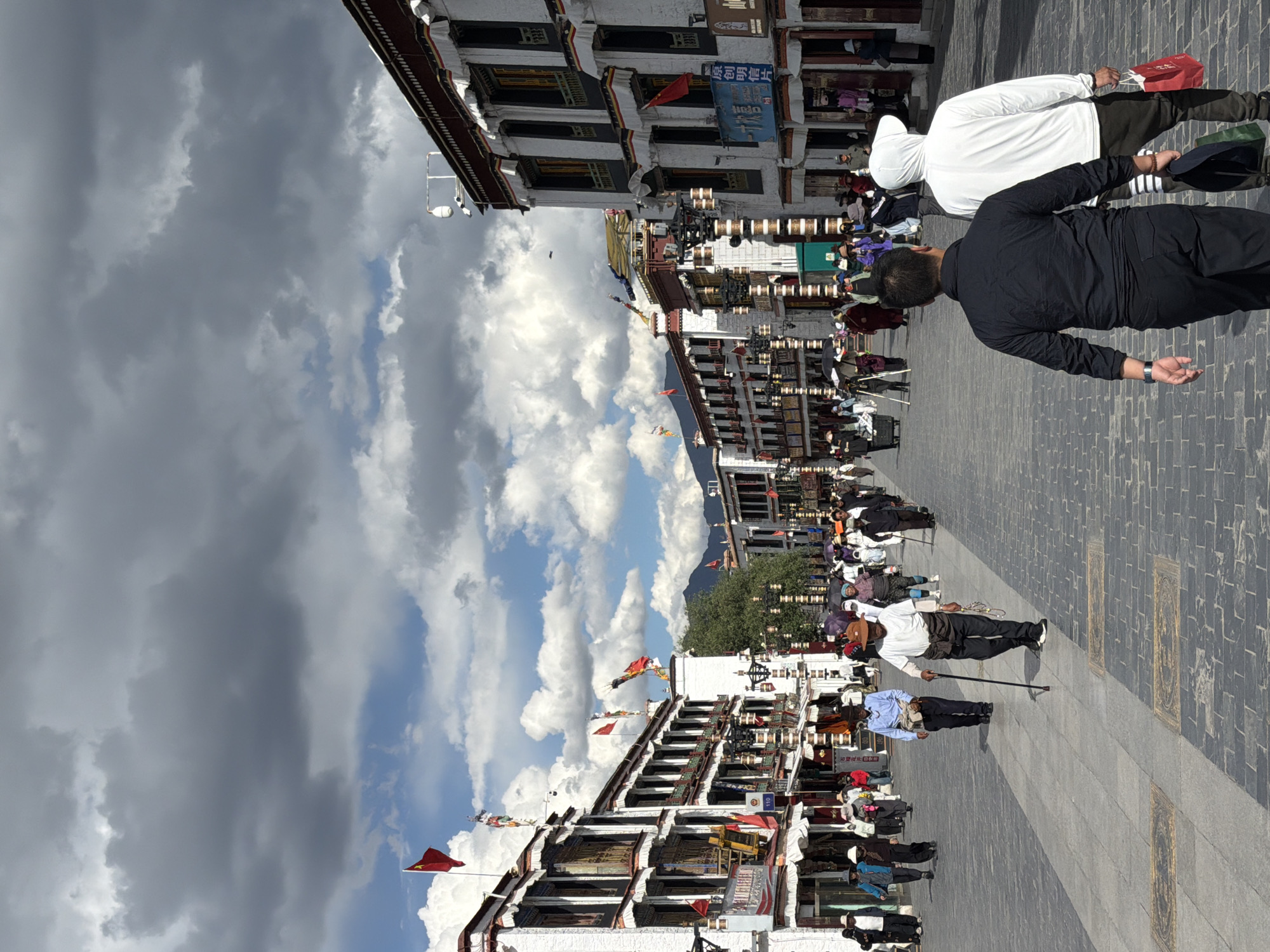 The image displays a bustling street scene within a traditional architectural setting, likely in Chengguan District, China, with a prominent mountainous backdrop. The perspective looks down a wide, paved street.
The sky is a striking blend of bright blue patches with fluffy white clouds and expansive areas of dark, heavy grey clouds, indicating variable weather conditions. Majestic mountains are visible in the distance, framed by the street and buildings.
In the foreground, two men walk away from the viewer on the paved street. One is dressed in a black long-sleeved shirt and black pants, while the other wears a white hooded top and dark pants, carrying a red bag. The street surface features a mix of light grey flagstones and darker brick-like paving, with rectangular golden-hued designs embedded at intervals.
Both sides of the street are lined with multi-story traditional buildings characterized by white walls, dark window frames, and ornate architectural elements. Many people are visible along the sides of the street, seated on the ground, some possibly engaged in prayer, prostration, or commerce. Further down the street, a man with a cane can be seen among the crowd.
Numerous red flags are displayed from poles and buildings, some clearly identifiable as Chinese national flags with yellow stars. A blue sign on one building prominently displays the text "明明电脑维修" (Mingming Computer Repair). A smaller red flag seems to bear the number "110".
The scene portrays a lively daytime atmosphere, capturing daily life, traditional architecture, and cultural activities within a scenic and dynamic environment.