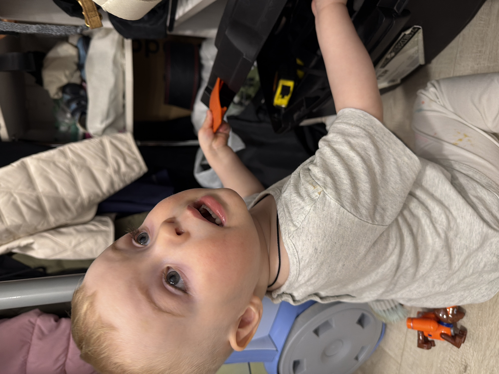 A young child with light skin and blonde hair is playing with objects in a cluttered storage area. The child is wearing a light grey t-shirt and white pants with stains. The child is holding an orange tool or part of a toy. The background contains a variety of items including clothes, bags, and a blue vacuum cleaner. The photo was not necessarily taken in Votkinsk, Russia, since there is no evidence for this.