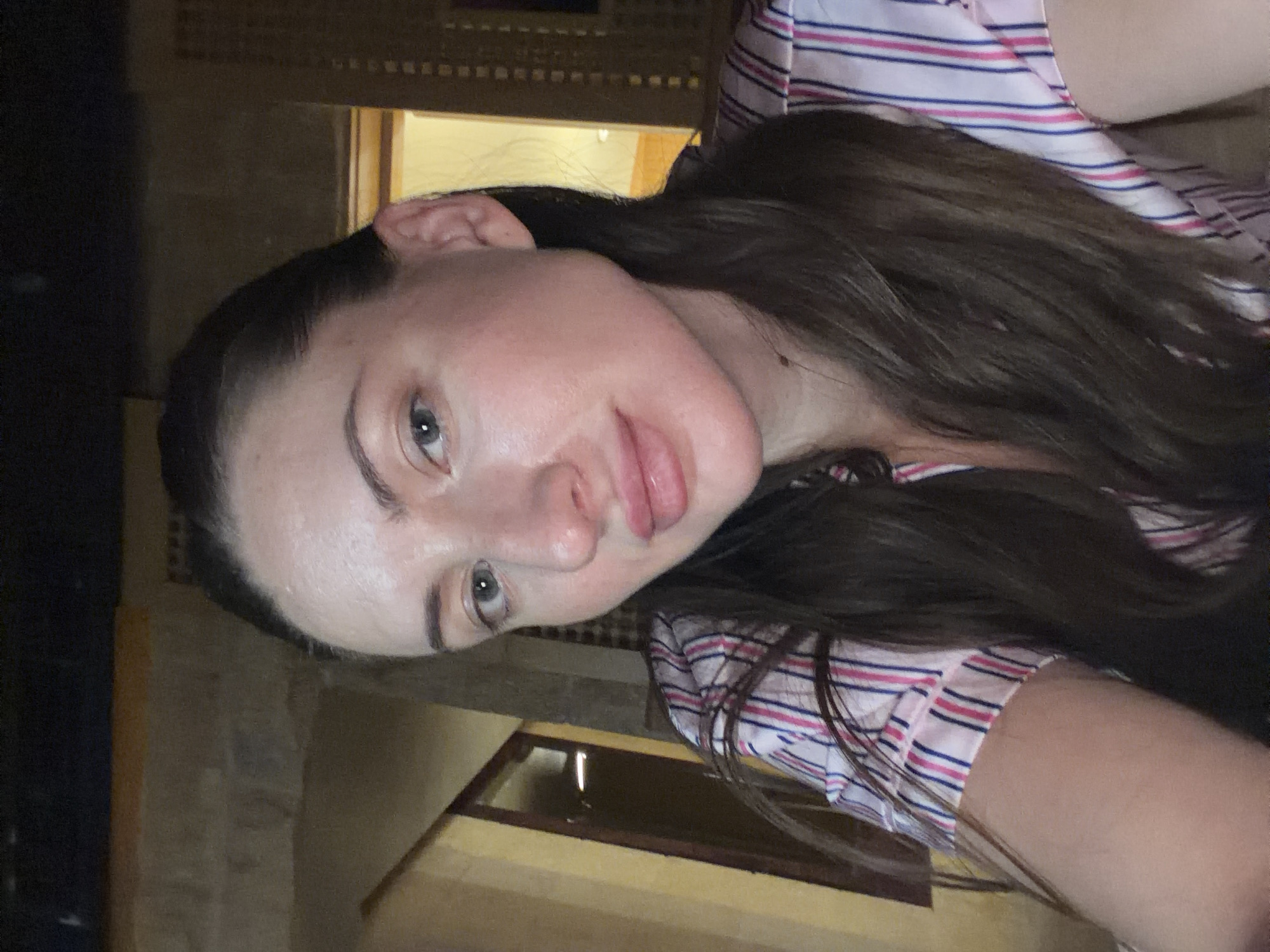 A medium shot captures a young woman with long, dark brown hair pulled back, exposing her forehead and ears. She is looking directly at the camera with a neutral expression. Her skin appears slightly dewy, and her lips are full and slightly parted. She is wearing a pink, white, and blue striped pajama top.
The background is dimly lit, suggesting an indoor setting at night. To the left, a portion of a yellow wall with a dark-framed doorway or window is visible. To the right, there is a glimpse of a wooden latticework or screen structure, and beyond that, a brightly lit doorway or opening. The overall impression is that the woman is in a hotel room or guesthouse, potentially in the city of Dahab, Egypt, given the architectural hints of latticework common in that region.