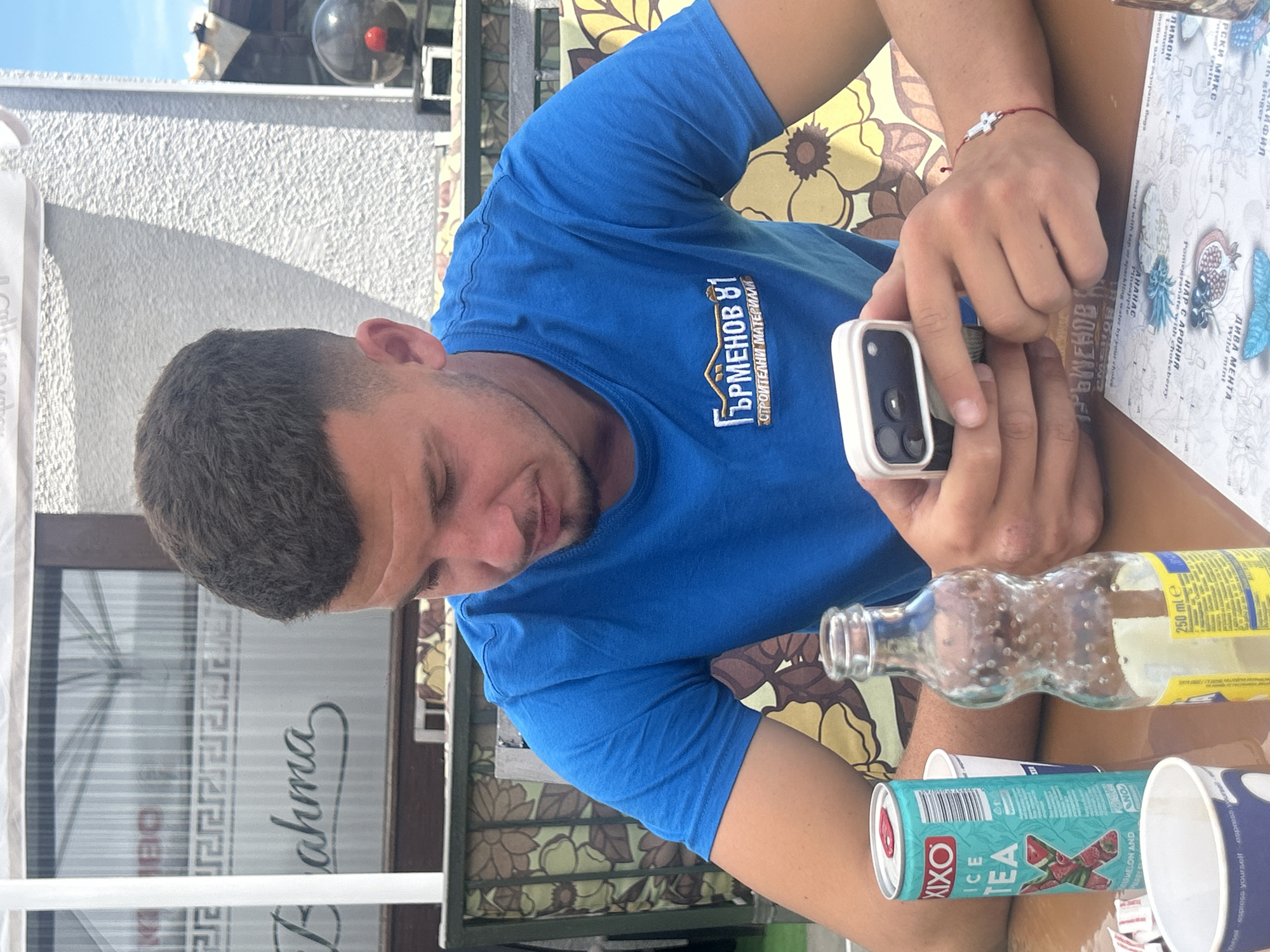 An overhead shot captures a man seated outdoors, engrossed in his white smartphone. The bright, sunny conditions are evident from the harsh shadows cast and the clear sky visible at the top left. The man, with short dark hair and a light beard, wears a blue t-shirt with the Cyrillic text "ГЪРМЕНОВ 81" (Garmenov 81) and "СТРОИТЕЛНИ МАТЕРИАЛИ" (Building Materials) printed on it. On his right wrist, he wears a red string bracelet adorned with a silver cross.
He is positioned at what appears to be an outdoor cafe or restaurant in Bania, Bulgaria, indicated by the Cyrillic text and general setting. A patterned fabric, possibly a cushion or tablecloth with a yellow and brown floral design, is visible beneath him. On the table or his lap, several items are placed: a clear glass bottle marked "250 ml e," a green and pink can of "XIXO ICE TEA WATERMELON AND," and two paper cups, one bearing the phrase "express yourself." The table surface is covered with a white sheet featuring illustrations of various fruits and plants, alongside Cyrillic descriptions such as "ЛИМОН" (Lemon), "ДИВА МЕНТА" (Wild Mint), "НАР С АРОНИЯ" (Pomegranate with chokeberry), and "АНАНАС" (Pineapple), as well as "Served with tap or sparkling water by your choice." In the background, a textured white wall, a metal railing, and a window reflecting "Fahma" are visible. The man's attention is entirely on his phone, suggesting a moment of leisure or communication.