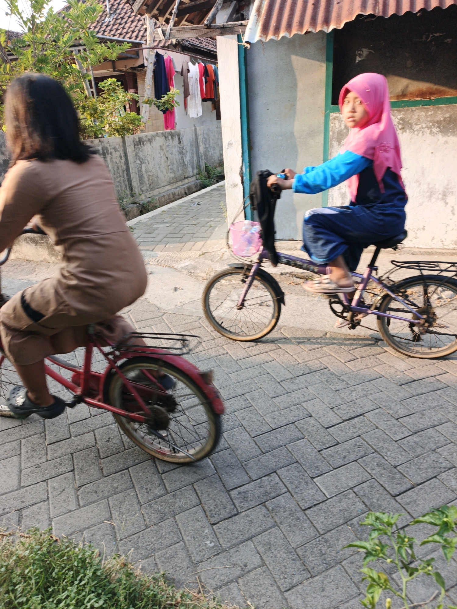 Two young girls are riding bicycles on a paved pathway in Ketegan, Indonesia. The girl in the foreground, with long dark hair, is wearing a brown outfit and riding a red bicycle. She is looking to the left and her body is blurred due to movement. The girl in the background is wearing a pink hijab and a blue and navy blue outfit. She is riding a purple bicycle with a pink basket and is holding a black object in her right hand. The bicycles are moving forward on a pathway made of interlocking grey pavers. Behind the pathway, there is a concrete wall, a fence, and a building with a corrugated metal roof. Clothes are hanging on a line against the building. Green foliage is visible on the left and in the lower right corner of the image. The lighting suggests it is daytime, with sunlight casting shadows.