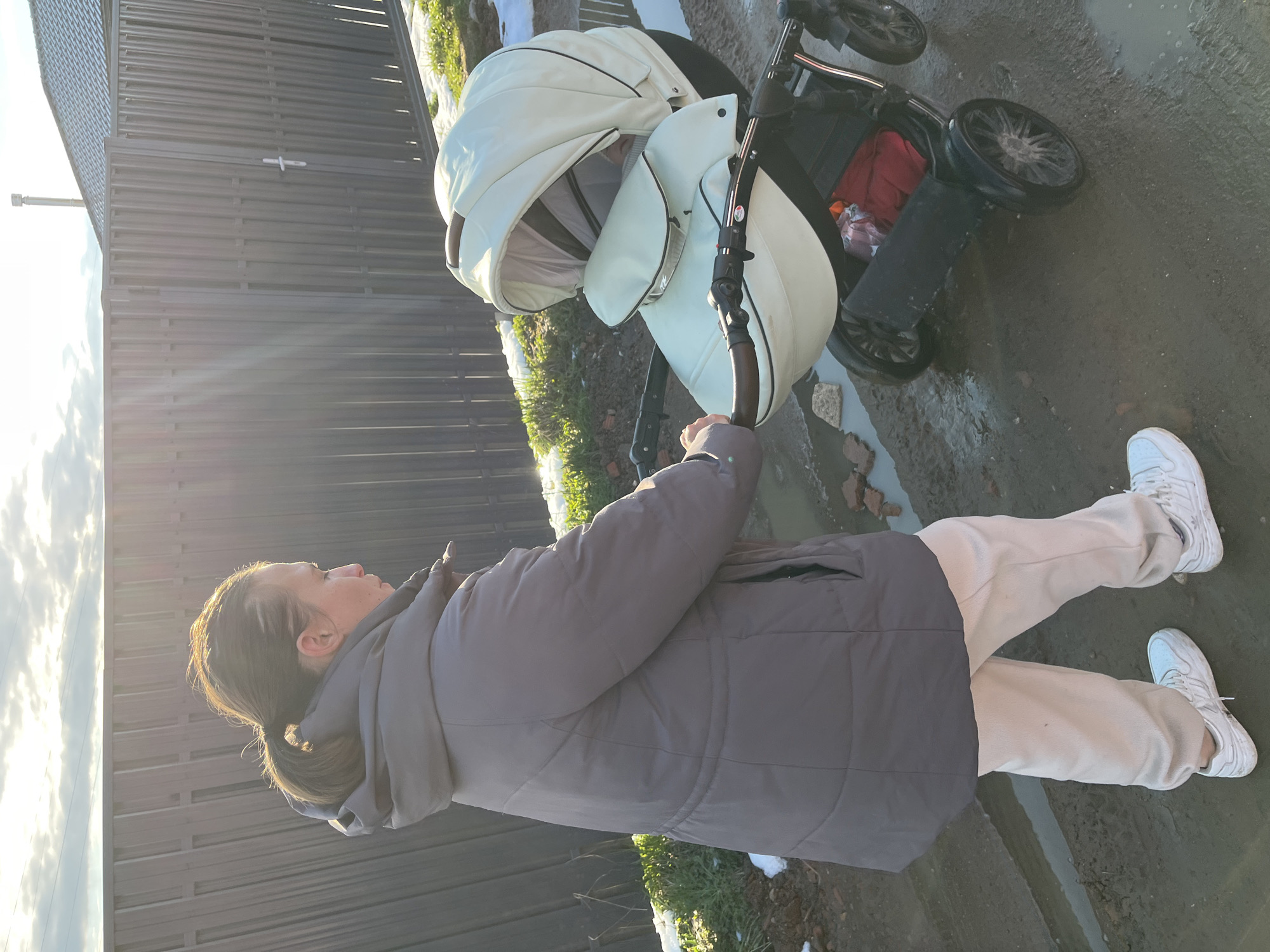 This image captures an outdoor scene, likely in Раздолье, Russia, featuring a woman and a baby stroller under bright sunlight. The woman, viewed from her left side, is wearing a dark grey puffer jacket, light-colored (possibly beige) pants, and white athletic sneakers. Her dark hair is tied back in a low ponytail. She is holding or pushing a cream-colored bassinet-style baby stroller with black accents and a brown-toned handle.
The setting appears to be a semi-rural or residential area. A prominent feature in the background is a tall, dark slatted fence. The ground is muddy, wet, and uneven, with visible patches of melting snow or ice, suggesting cold weather conditions. There are also scattered small pieces of brick or debris on the ground, and sparse winter vegetation. The strong sunlight casts bright glare and suggests either early morning or late afternoon. The activity depicted is a casual outdoor stroll, possibly with a baby, in a rugged outdoor environment. No visible text is present in the image.