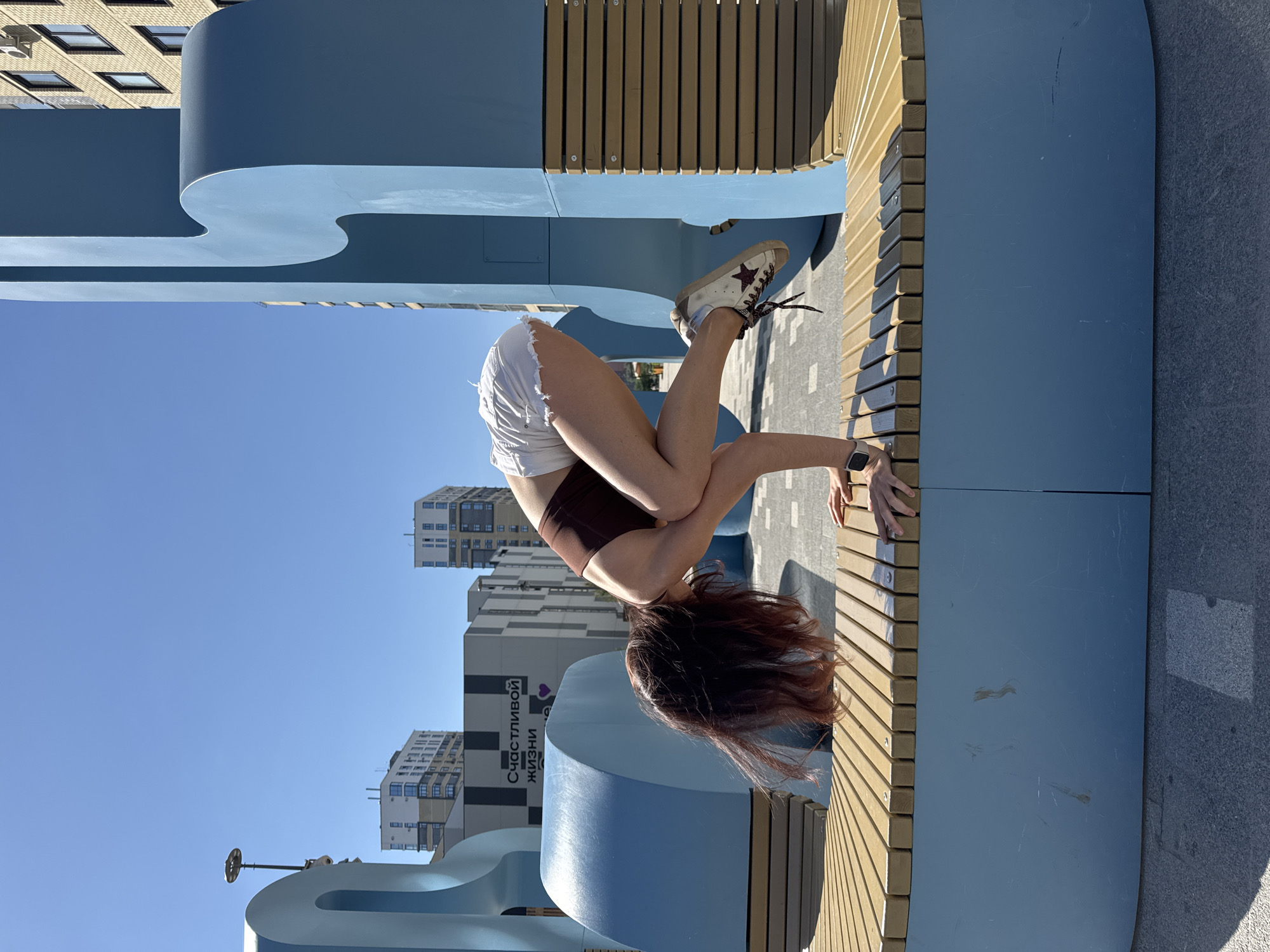 A woman with red hair and a brown sports bra and white shorts is bent over on a bench in an outdoor setting, possibly in the city of Tyumen, Russia. She is wearing sneakers with a star and her hands are on the bench. The bench has a blue base with wooden slats. Behind her are buildings, including one with a sign in Russian that includes the words "Счастливой жизни". The sky is blue and sunny.